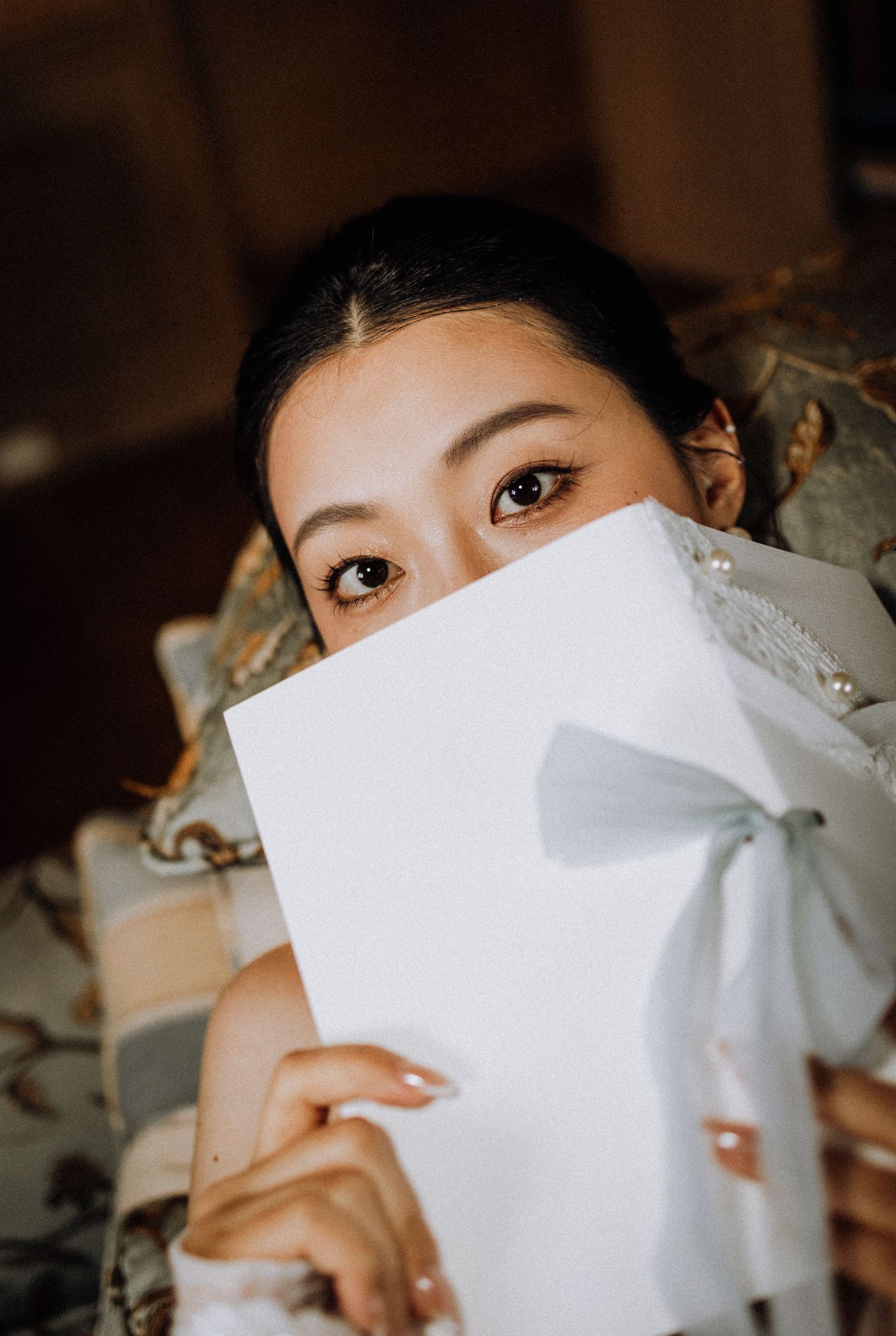 A young Asian woman playfully hides her face behind a white gift box with a ribbon, revealing only her eyes.