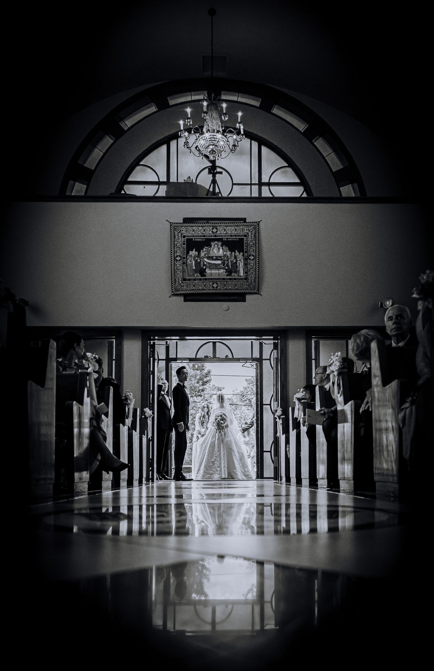 Black and white wedding photo of bride and groom silhouetted in church doorway with chandelier above.