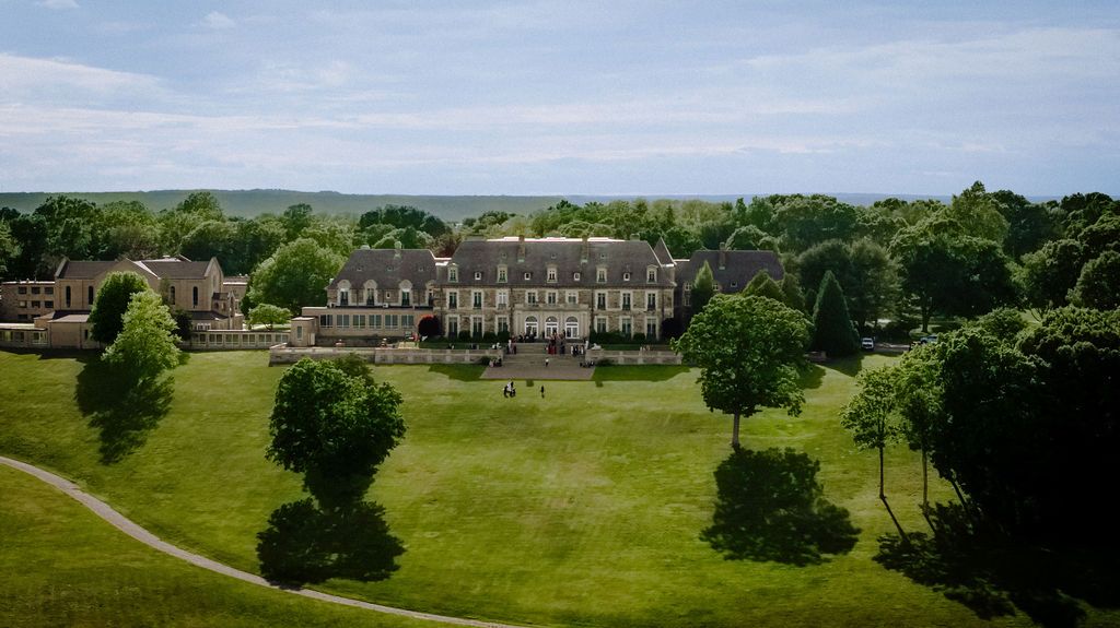 Aerial view of a grand historic mansion surrounded by lush green lawns and trees under a partly cloudy sky.