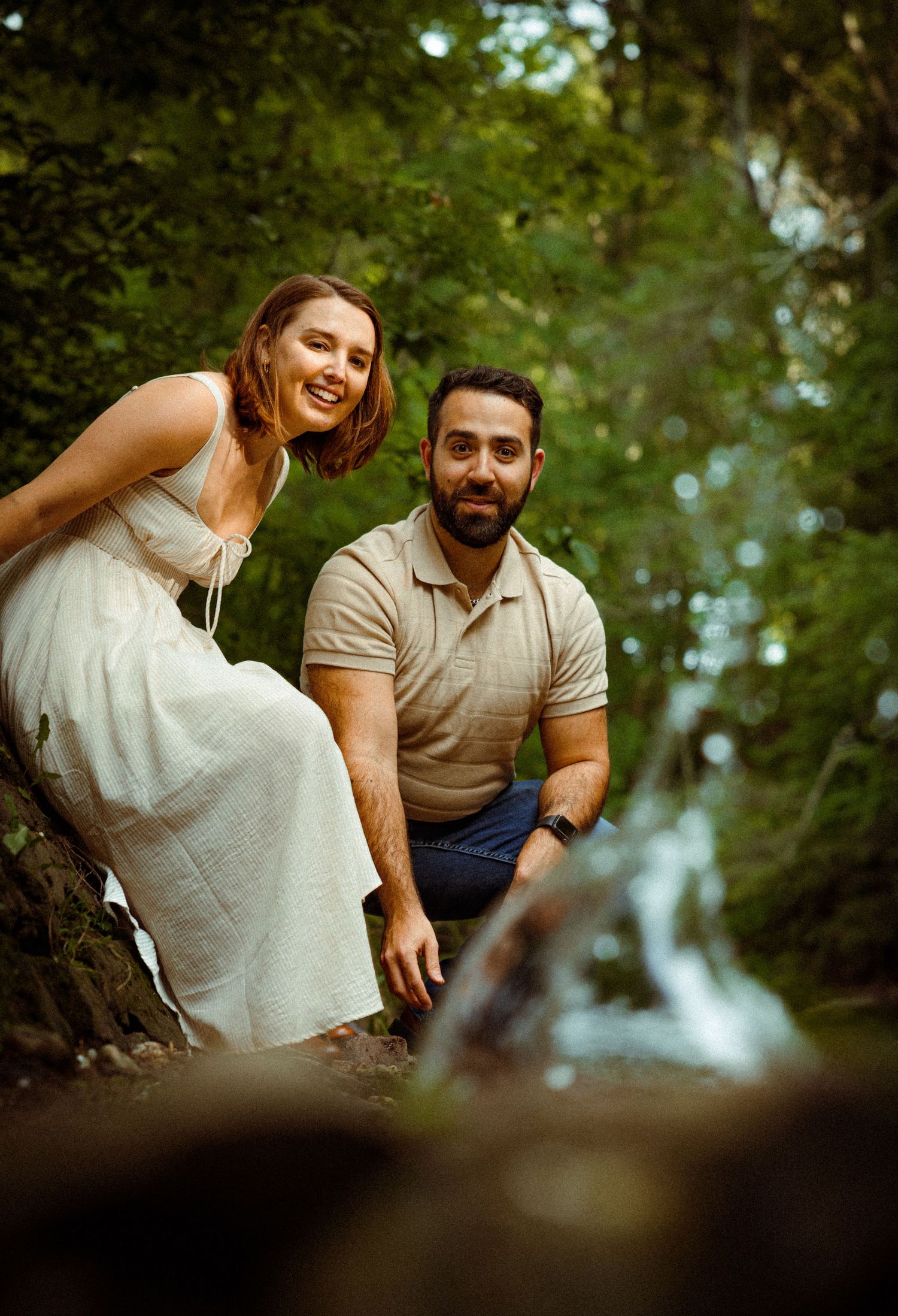 A couple poses together on a forest path with a soft blurred waterfall visible in the background.