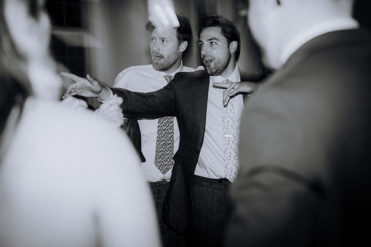 Two men in suits and ties share a lively moment on a wedding dance floor in black and white photo.