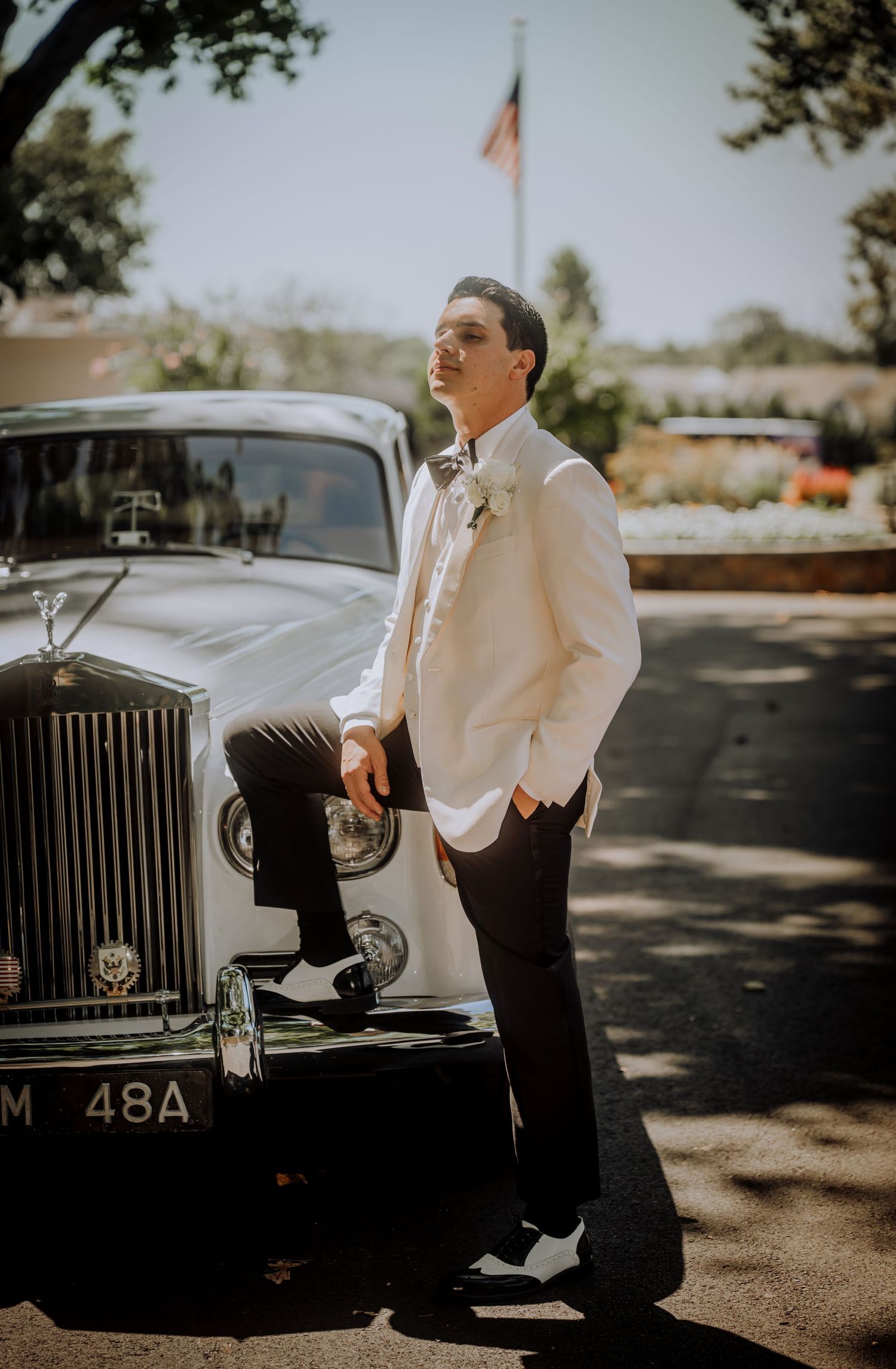 Groom in white tuxedo poses beside classic Rolls Royce under American flag.