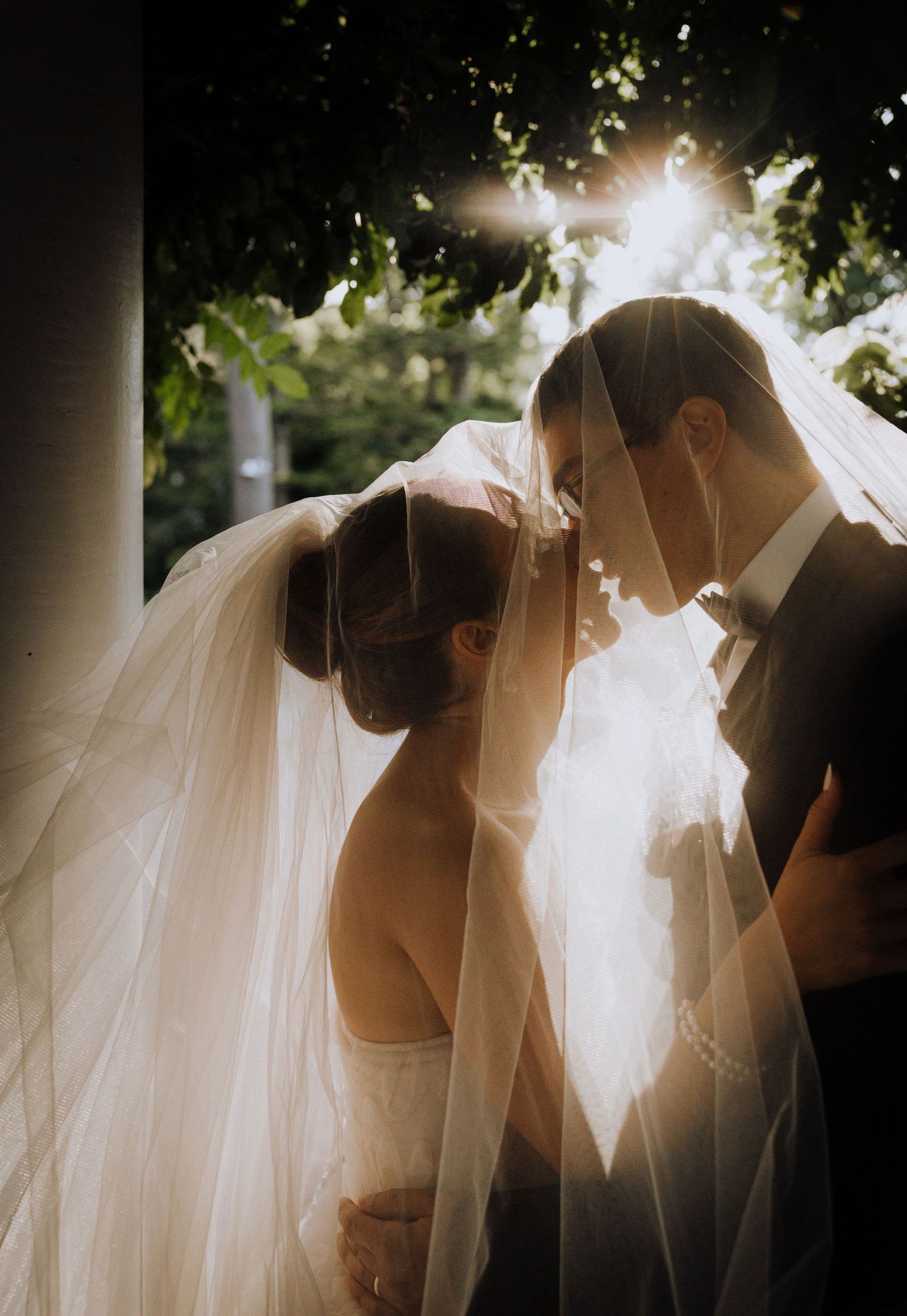 Bride and groom sharing an intimate moment under a wedding veil with sunlight filtering through trees.
