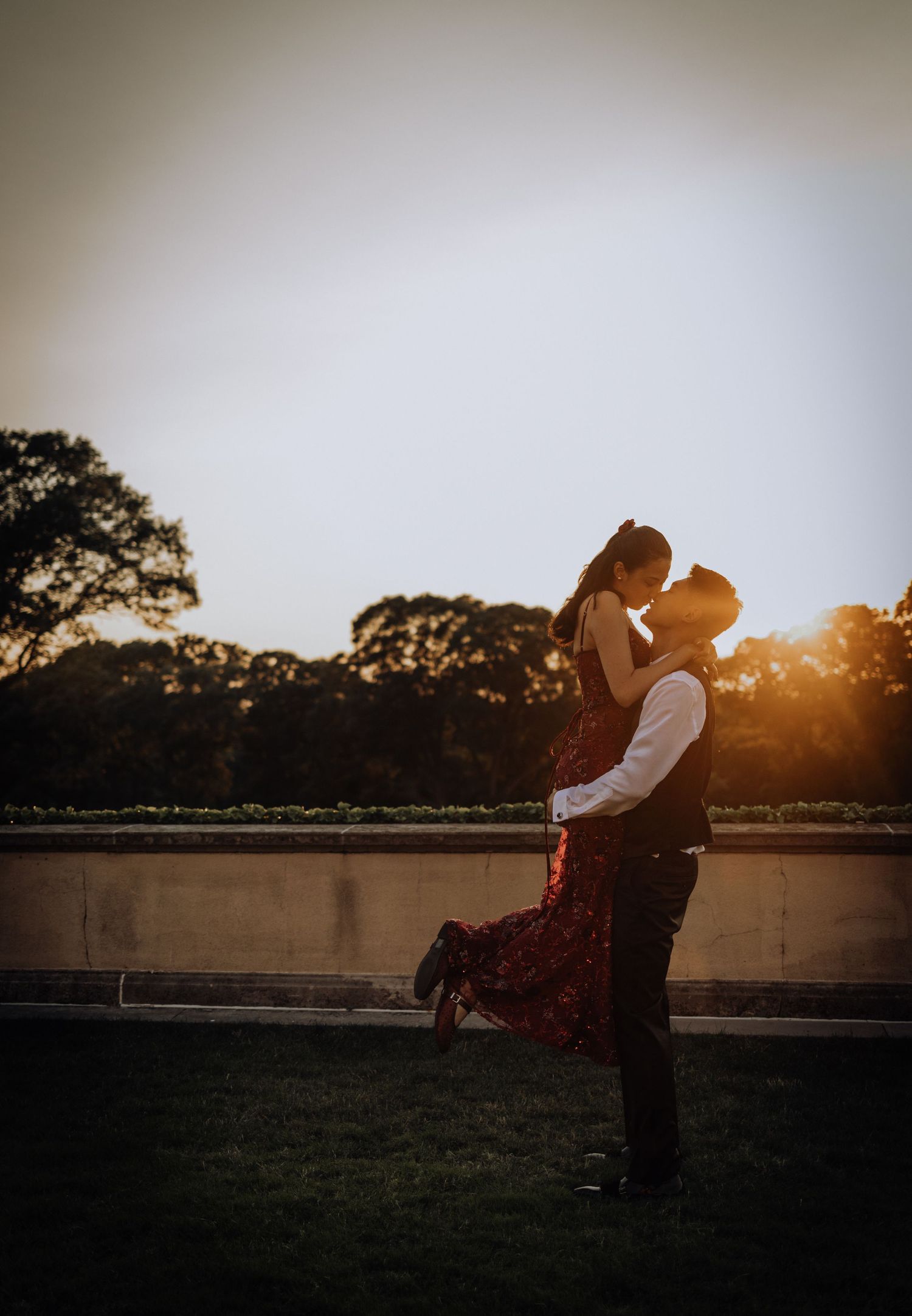 Couple sharing a romantic kiss at golden hour sunset, man lifting woman in red floral dress outdoors.