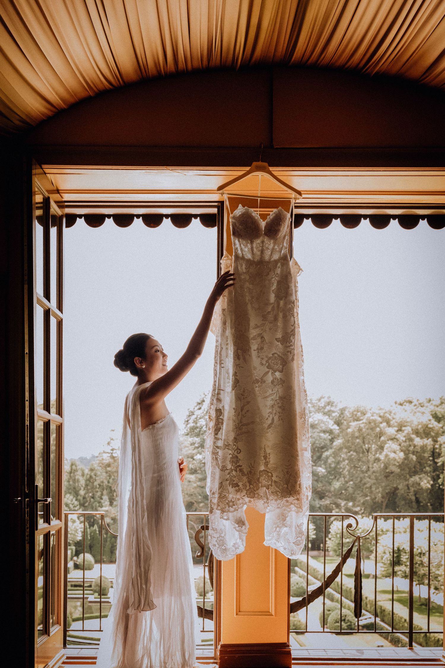 Bride in white robe reaches for lace wedding dress hanging on balcony door in elegant venue.