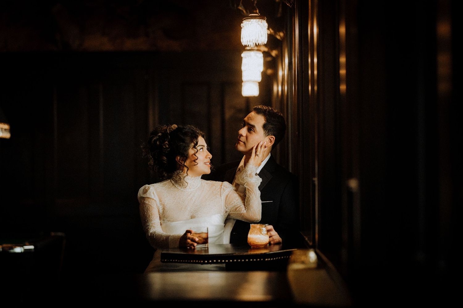 Romantic couple in wedding attire sharing an intimate moment at a dimly lit bar, bride touching grooms face tenderly.