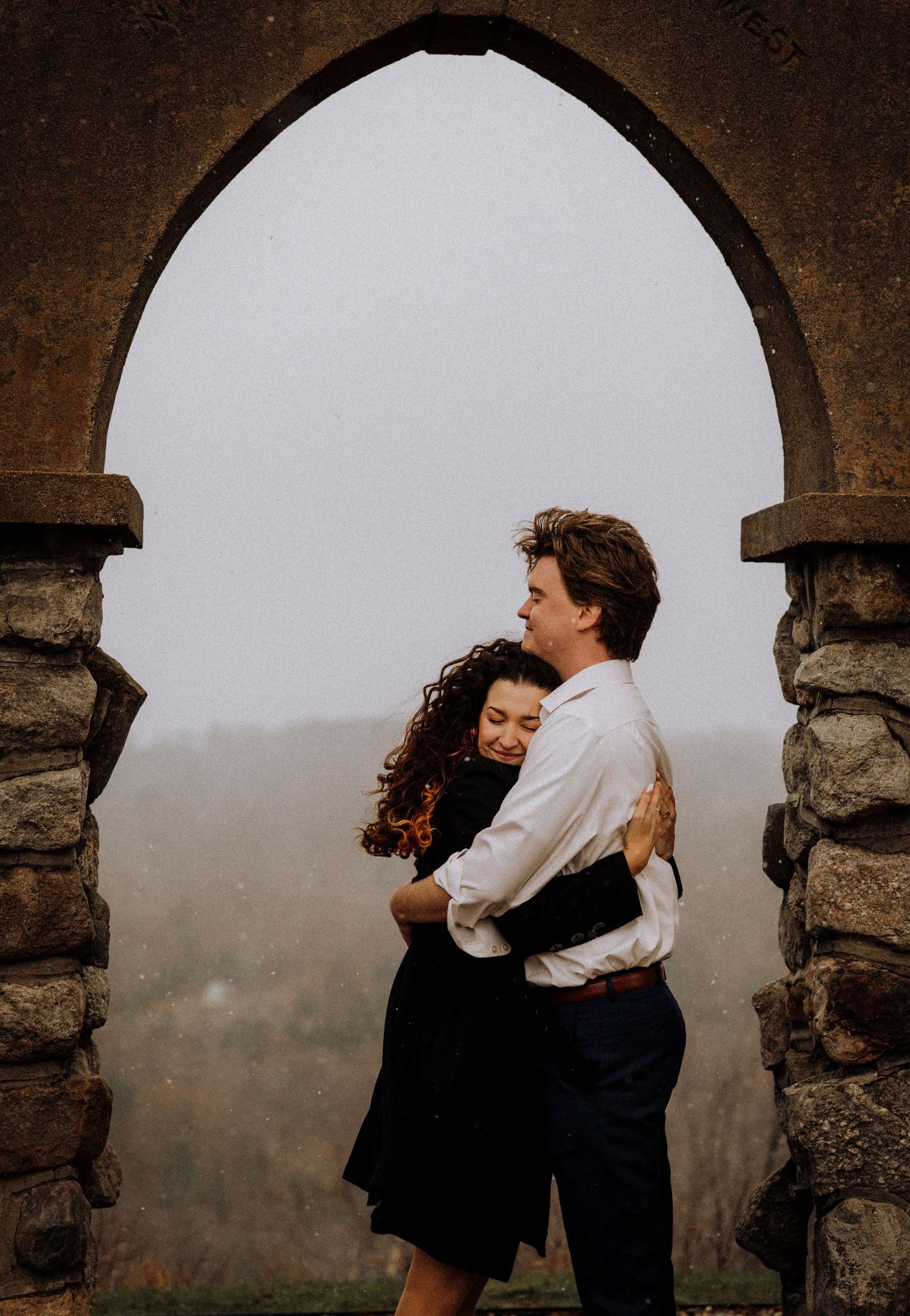 A couple embraces under stone archways against a misty mountain backdrop.
