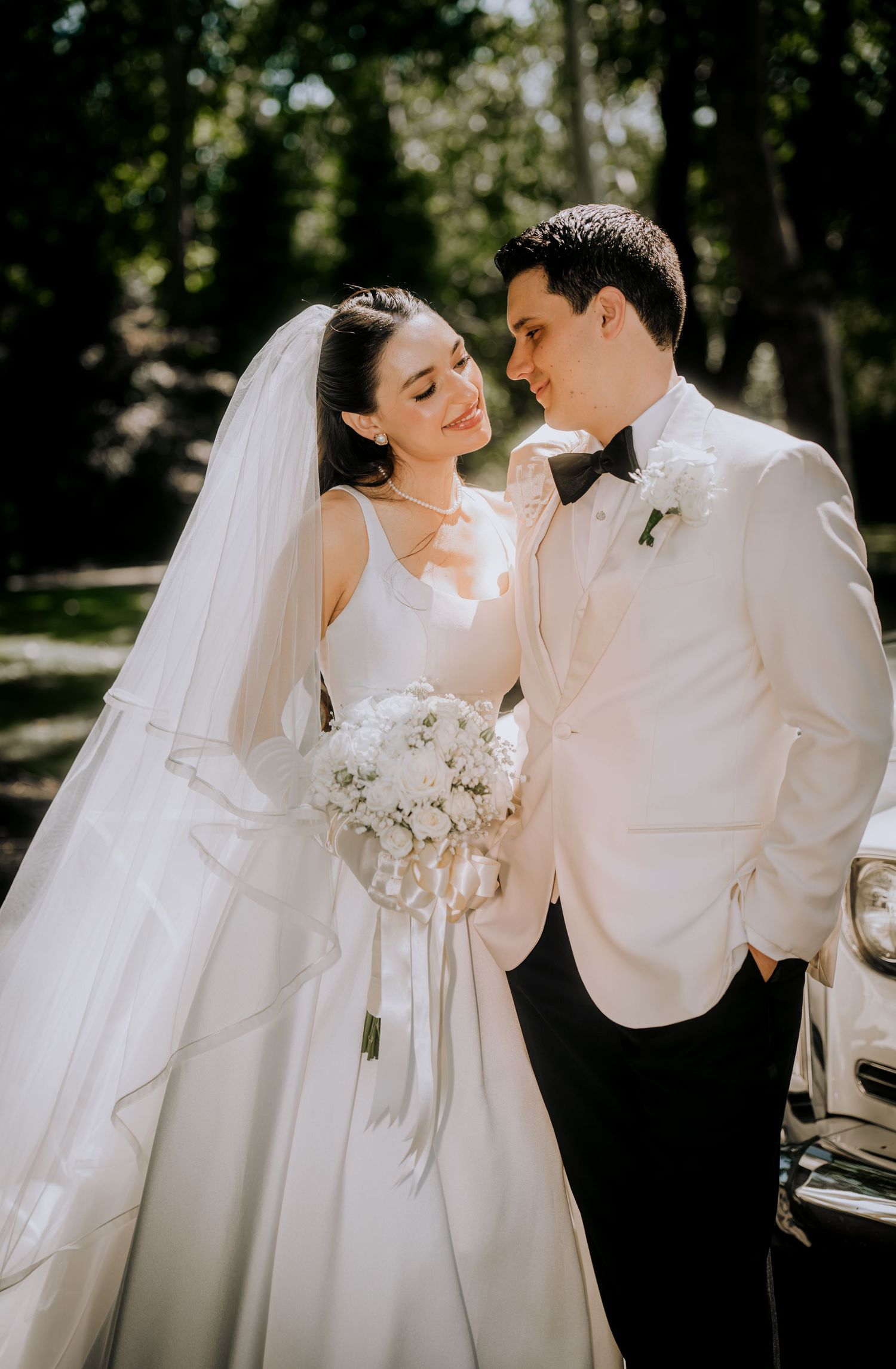 Elegant wedding couple in white attire pose together in a sun-dappled garden setting.