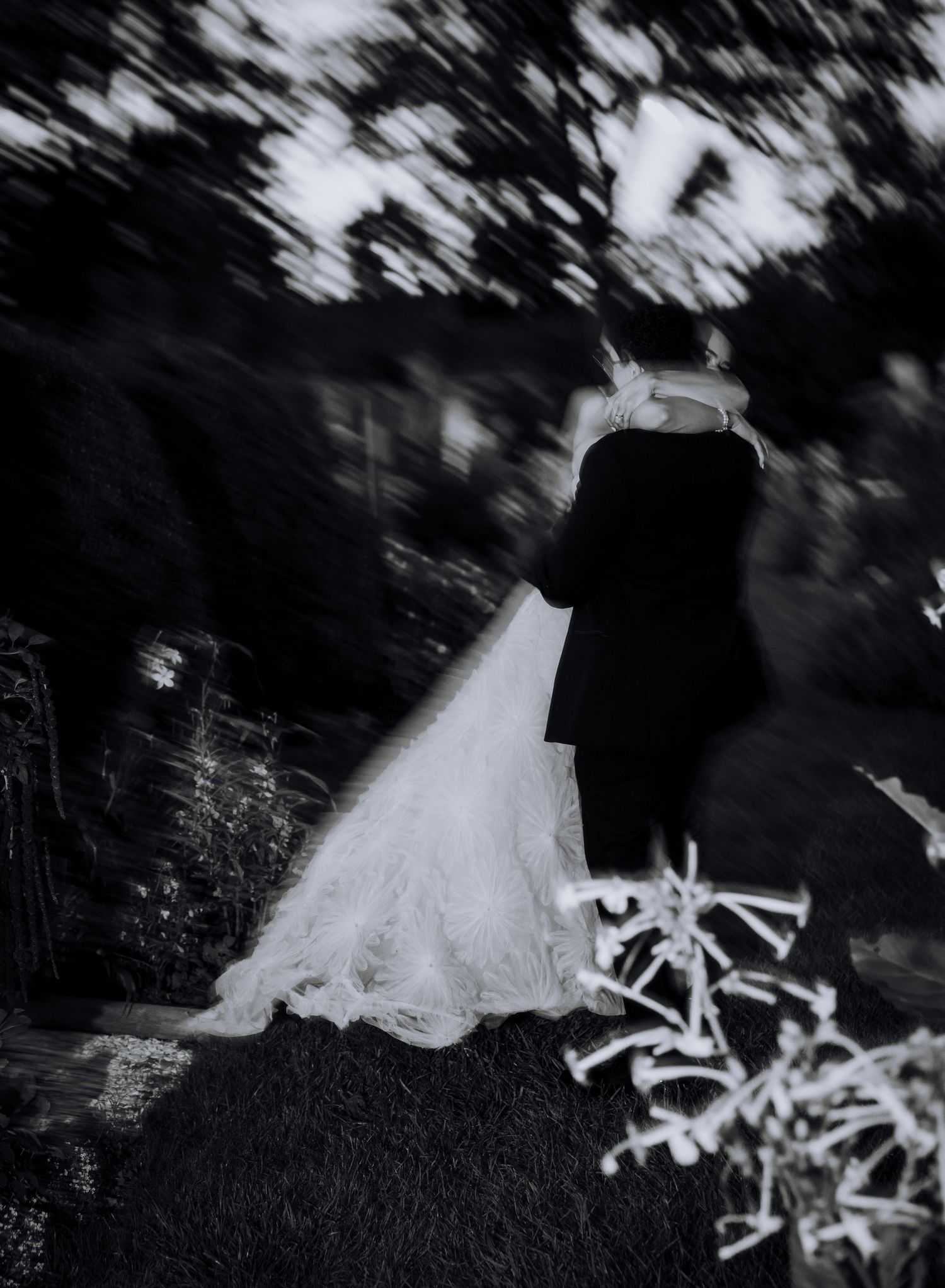 Black and white blurred photo of a bride and groom embracing outdoors, wedding dress train flowing on ground.