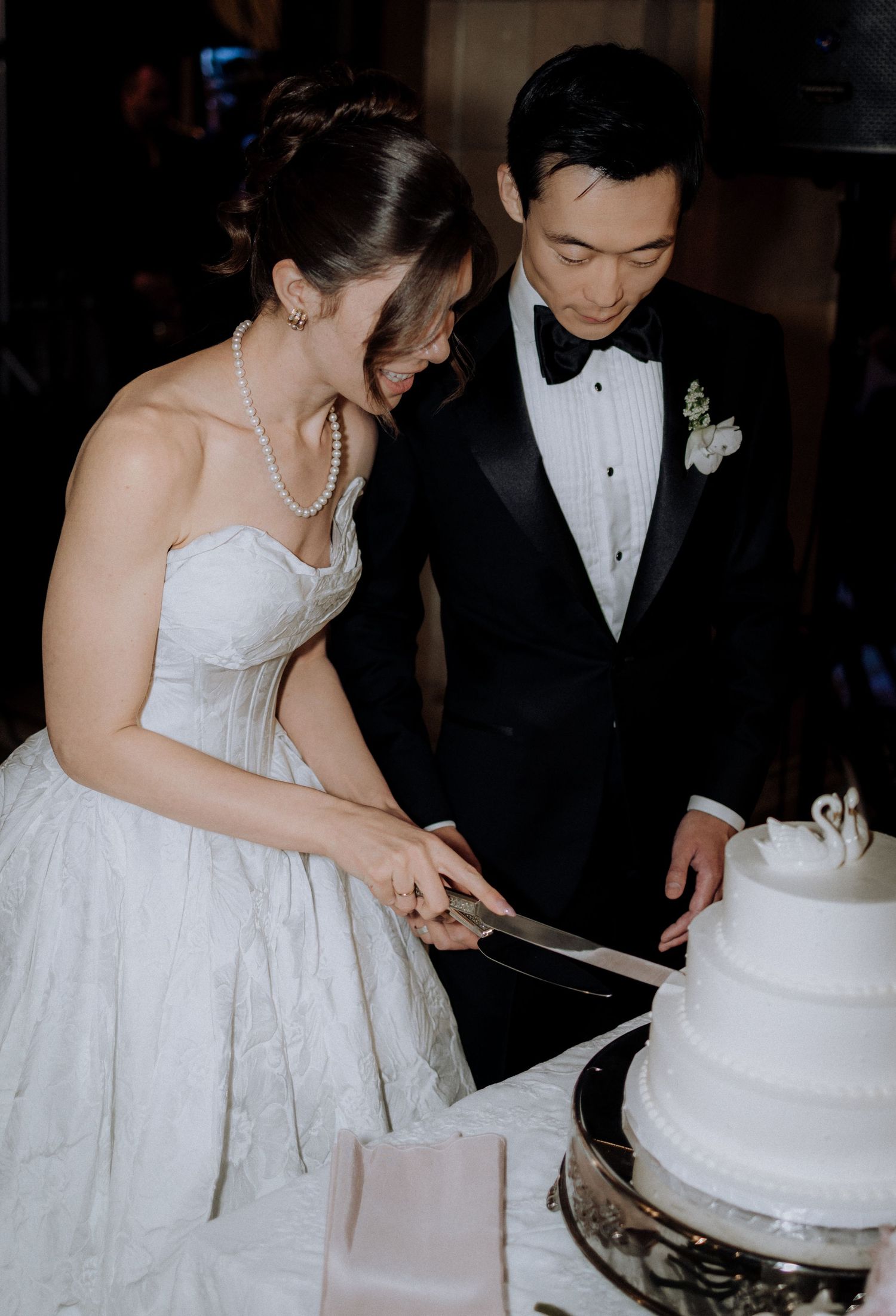 Bride and groom cutting white tiered wedding cake, bride in strapless gown with pearl necklace, groom in black tuxedo.