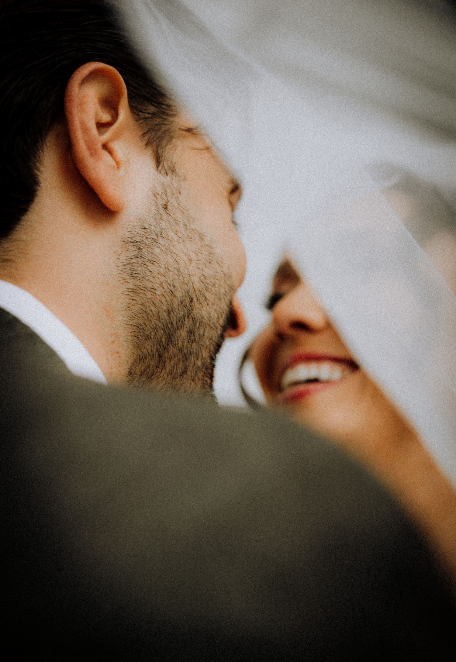 A bride and groom share an intimate moment under a white veil, smiling closely together on their wedding day.
