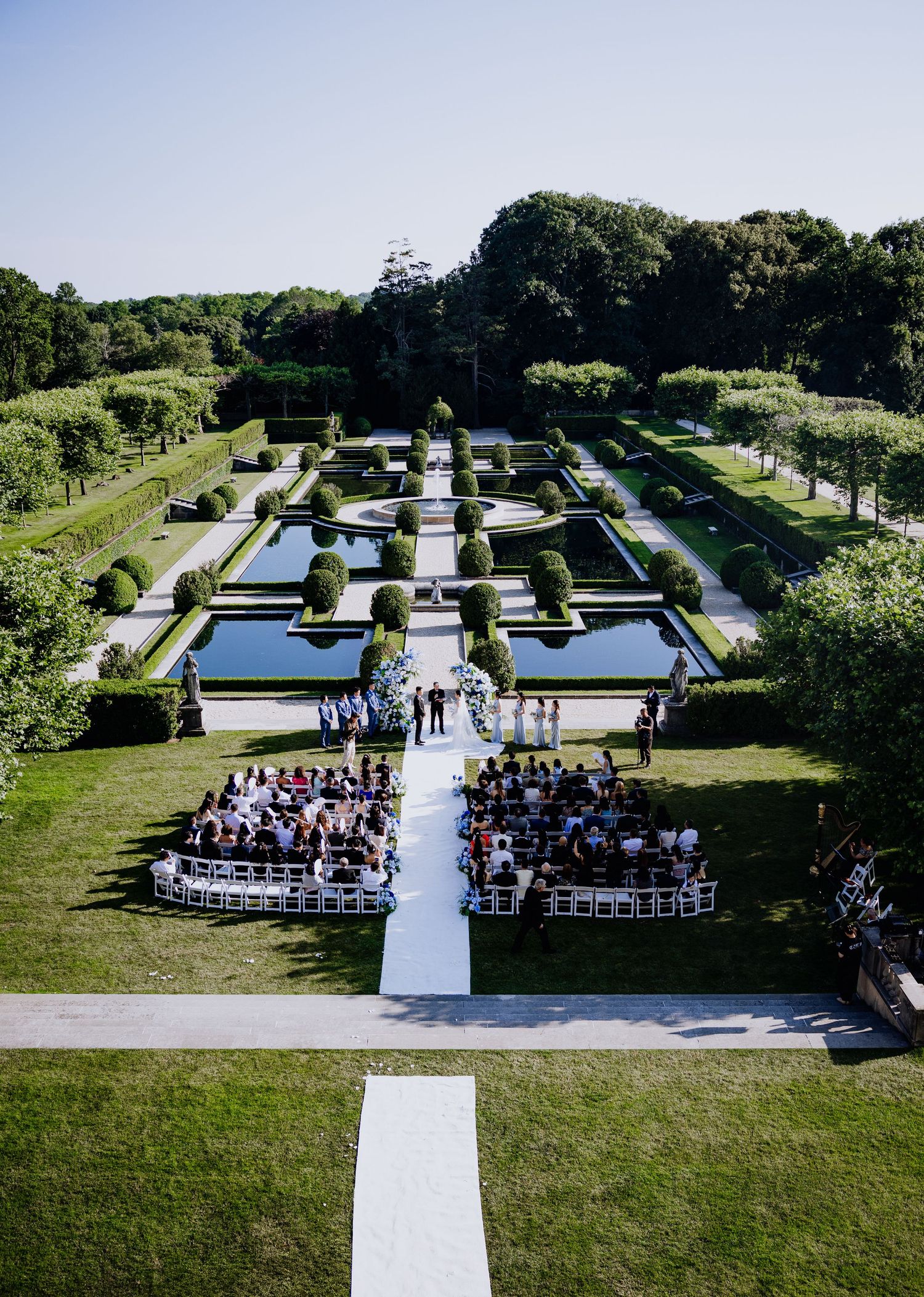 Aerial view of an elegant outdoor wedding ceremony in a formal garden with manicured hedges and reflecting pools.
