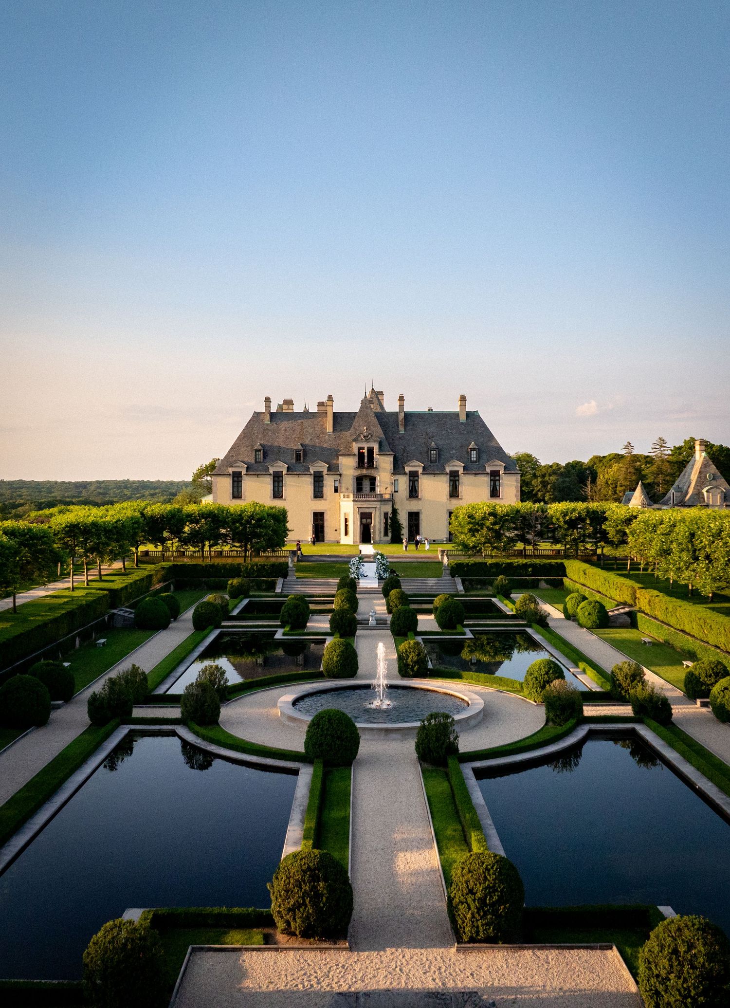 Aerial view of a grand French chateau with formal gardens, fountains, and manicured hedges under a clear blue sky.