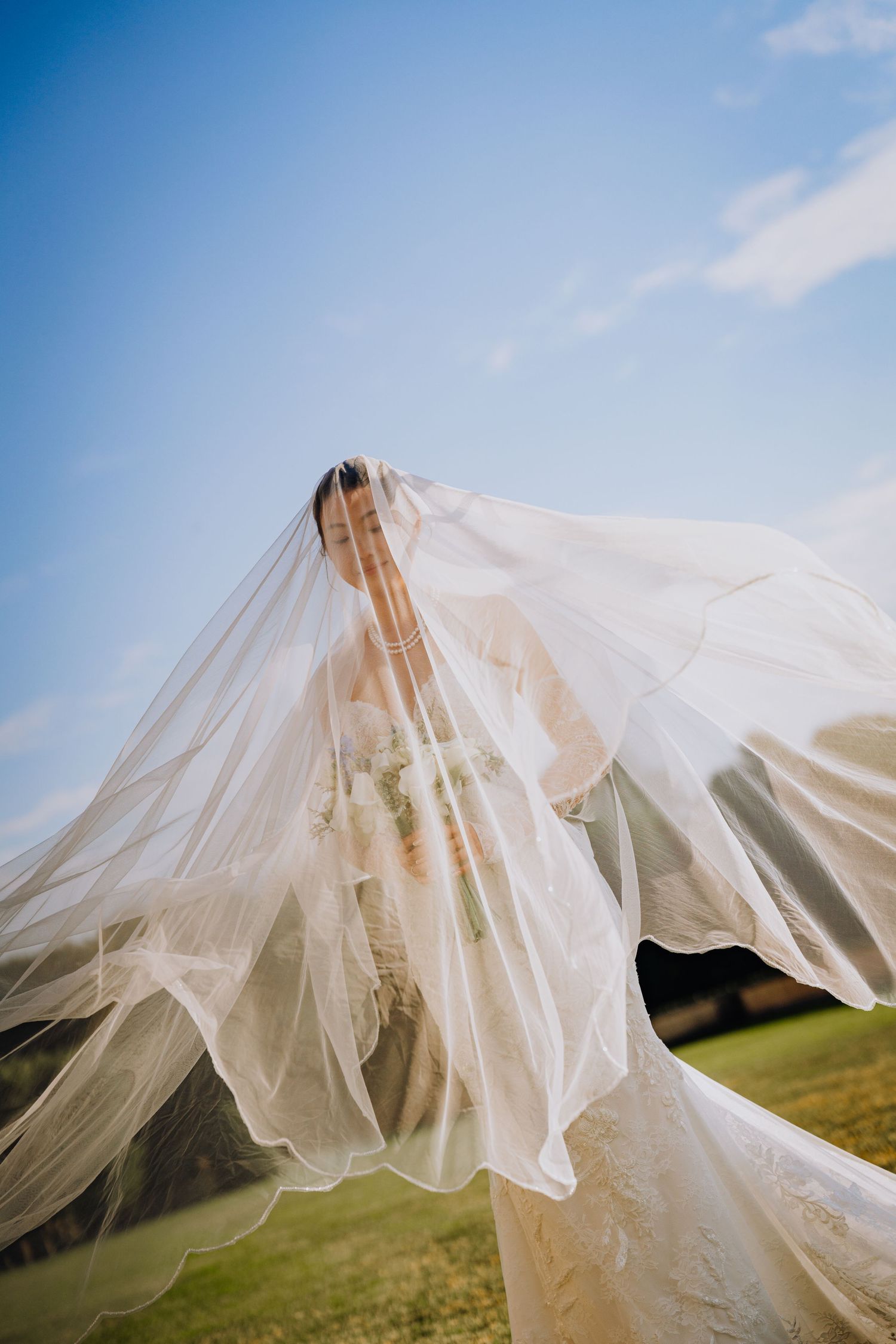 Bride in white lace gown with flowing veil billowing in wind against blue sky on green lawn.