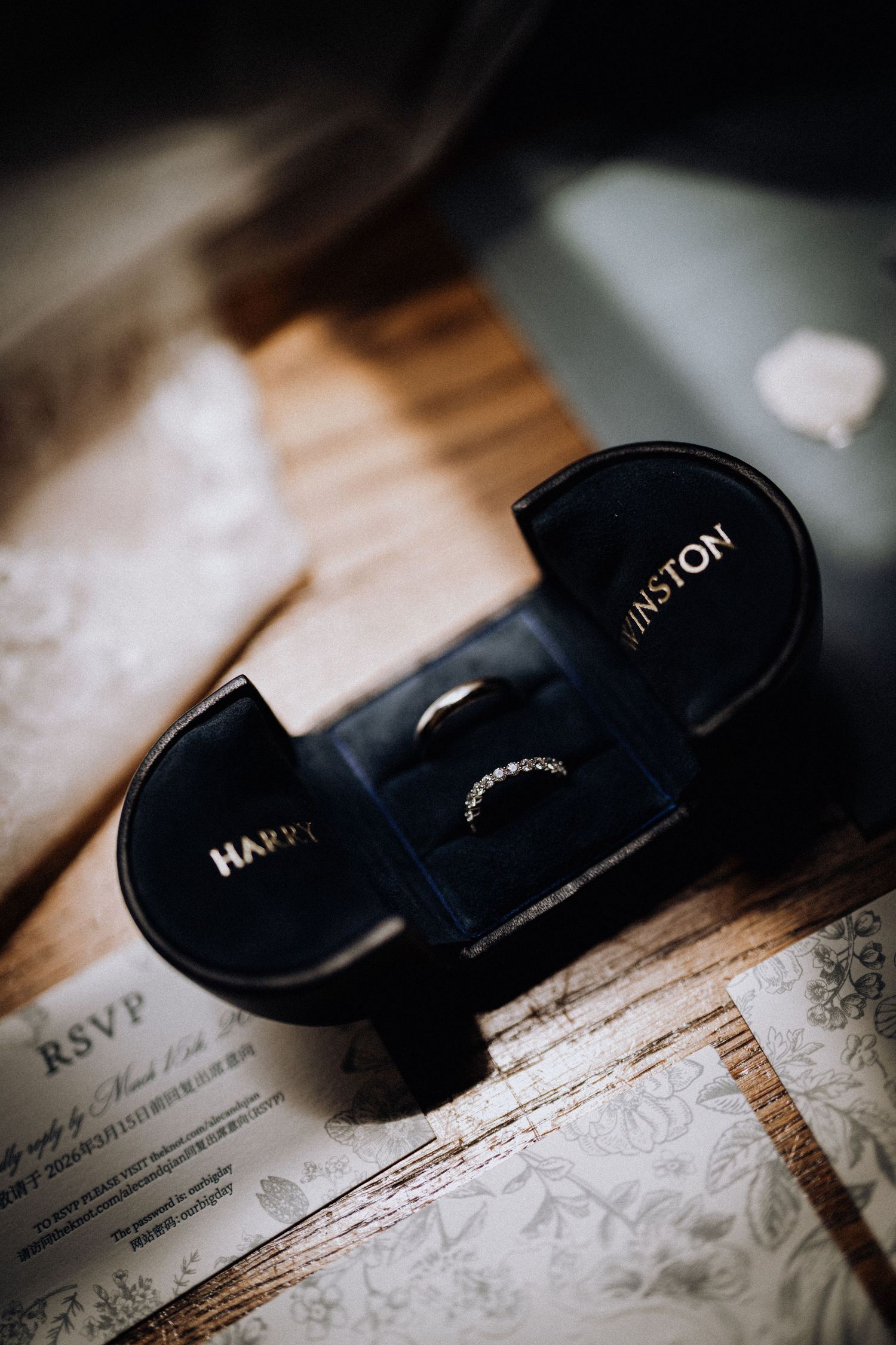 Two wedding bands displayed in a black Harry Winston ring box on a wooden surface with RSVP card.