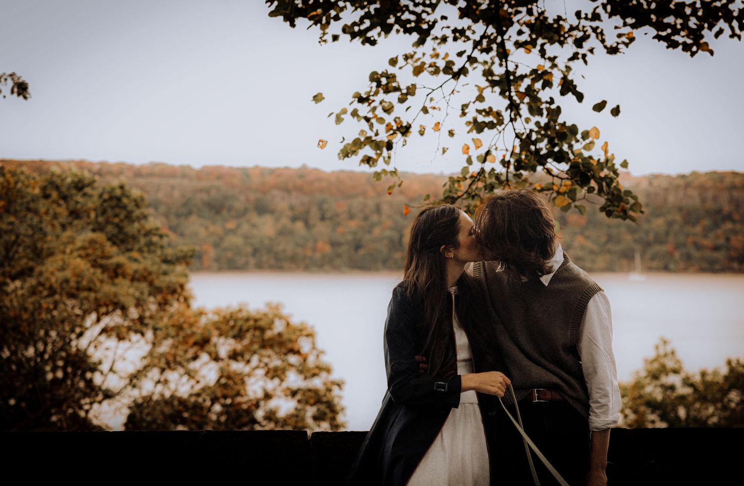 A couple embraces while overlooking an autumn lake surrounded by fall foliage.