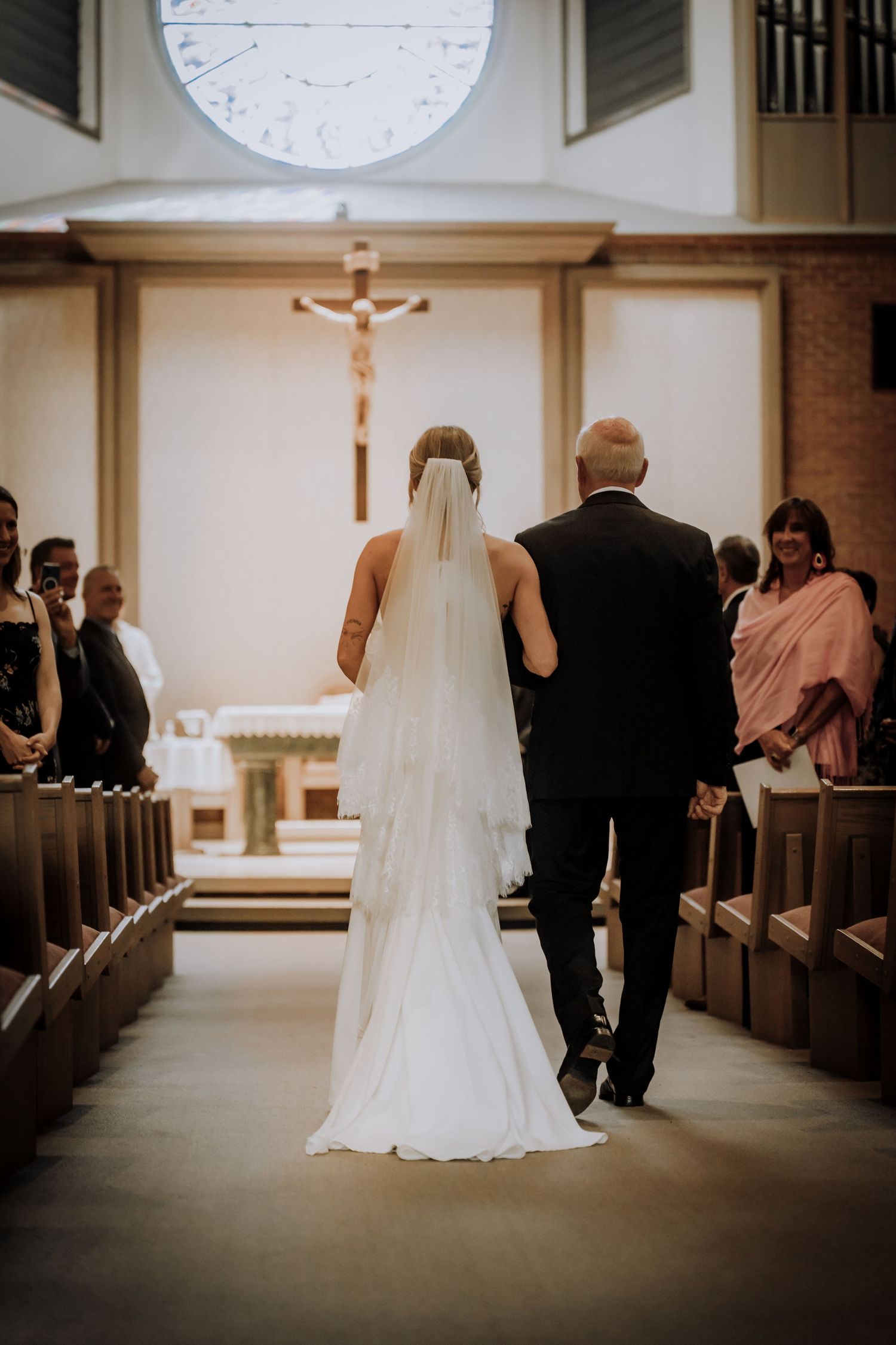 A bride walks down the church aisle with a long flowing veil beneath a crucifix.