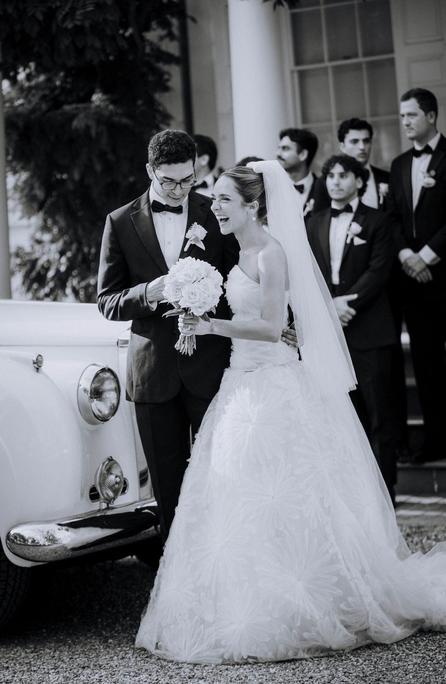 Black and white photo of a smiling bride in a white gown and groom in tuxedo beside a vintage car at their wedding.