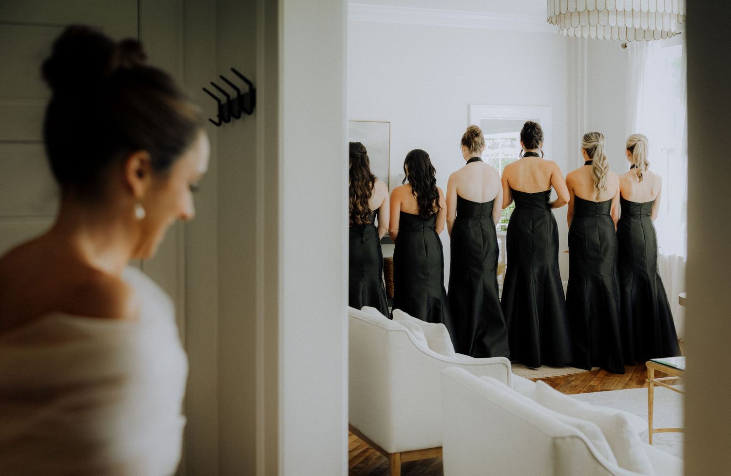Bride in white gown with bridesmaids in black dresses standing in elegant room during wedding preparation.