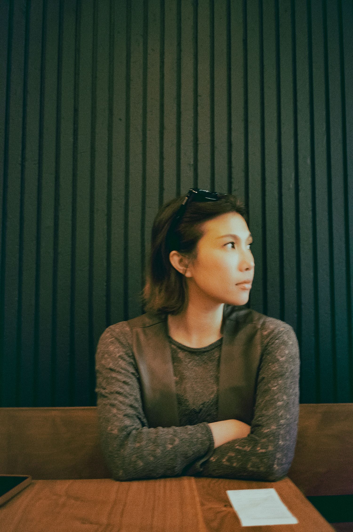 Young woman with sunglasses on head sits cross-armed at wooden table against dark slatted wall.