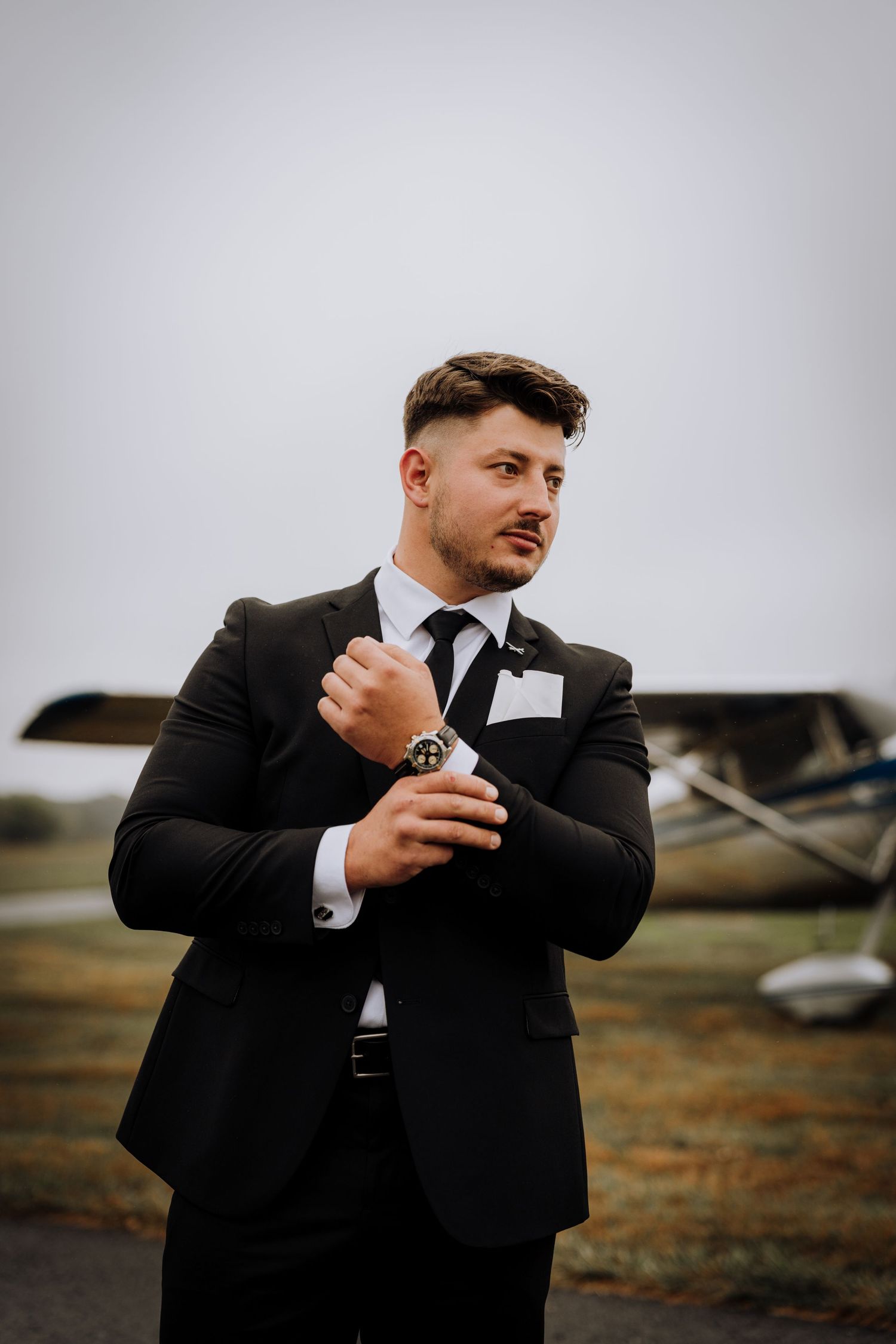 Groom adjusting his tie while standing in formal attire near an aircraft.