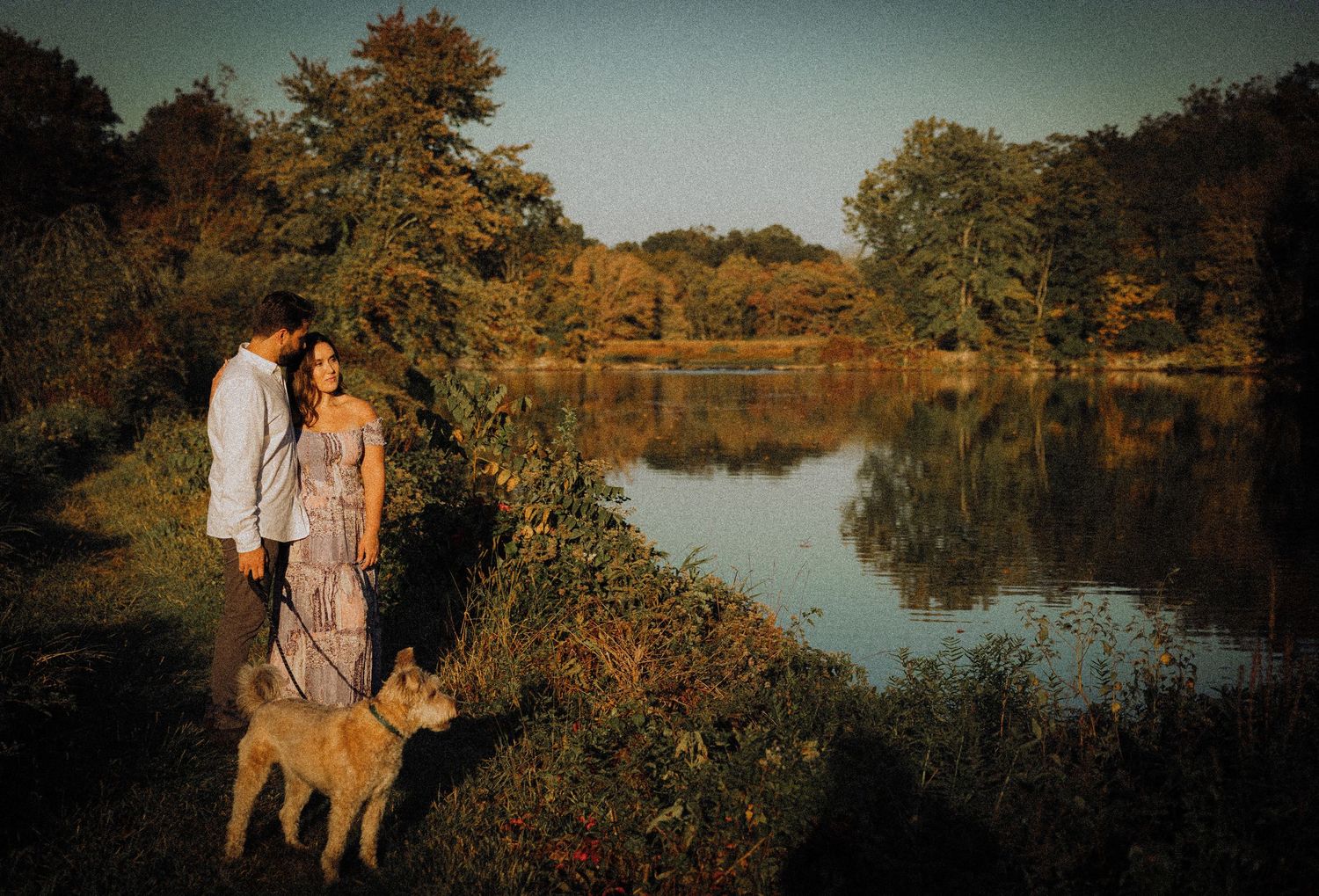 Couple with their dog standing by a serene lake during autumn sunset.