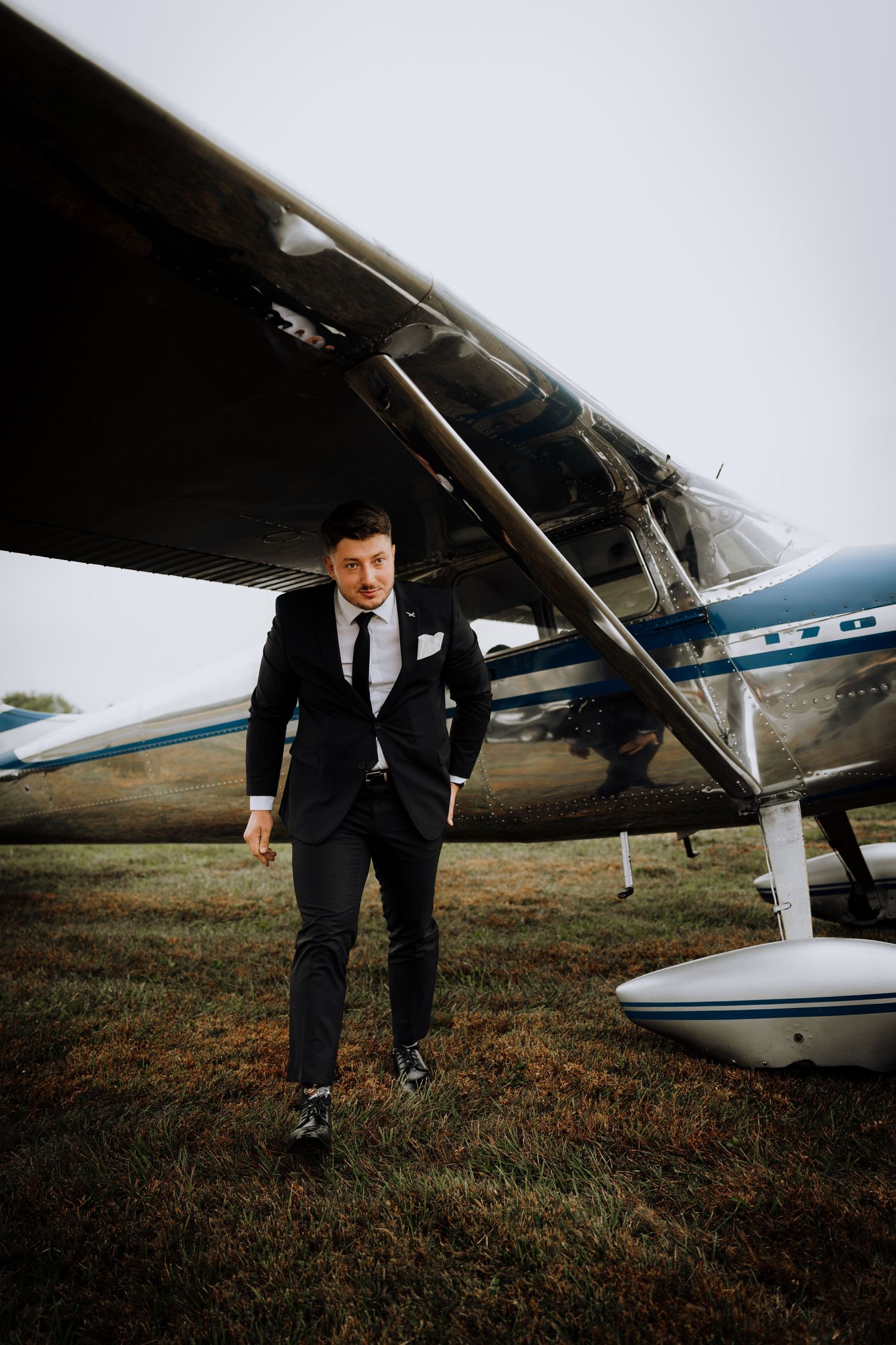 A groom in a black suit poses confidently next to a small aircraft.