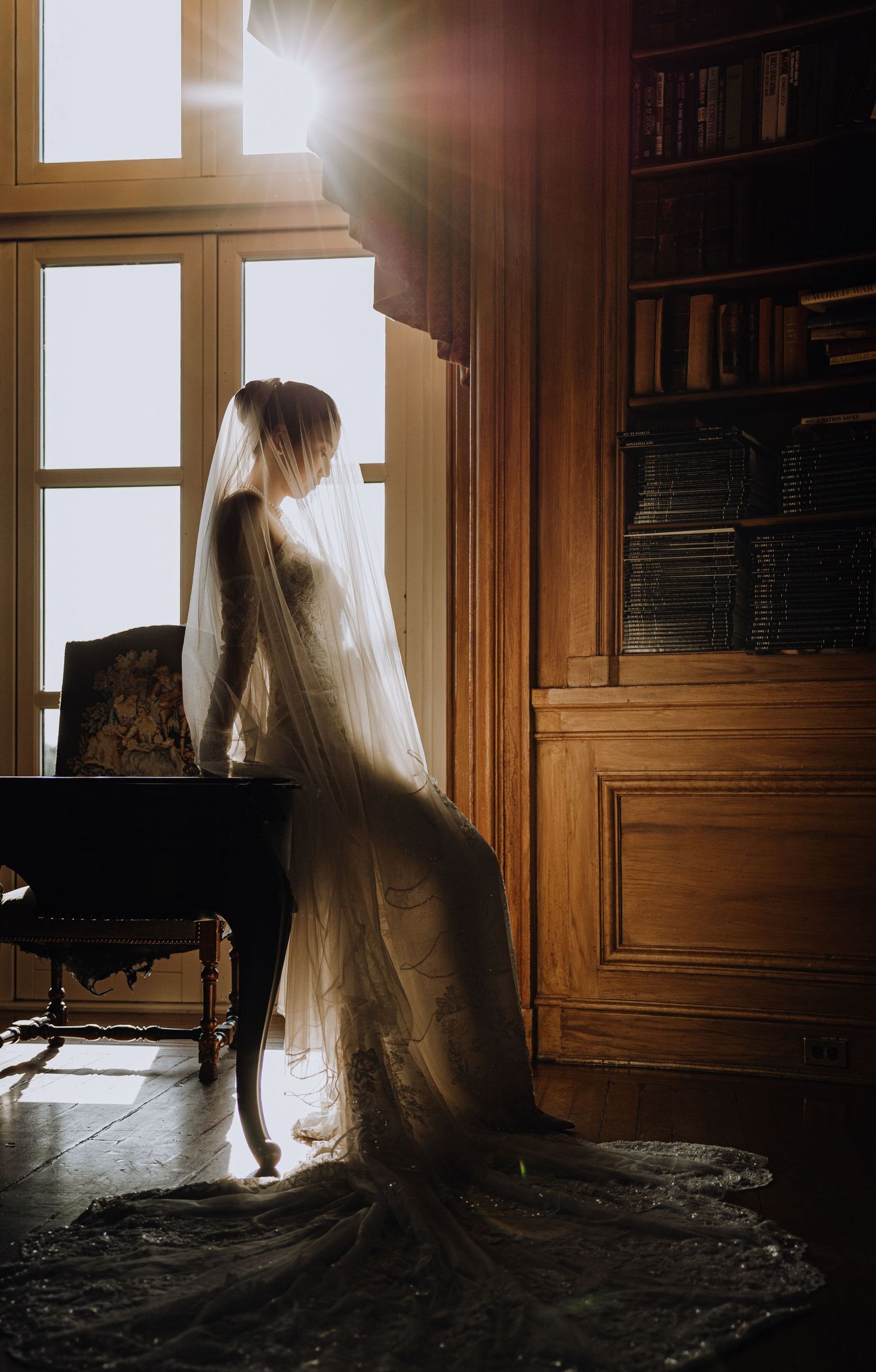 Bride in a flowing wedding gown and veil stands by a sunlit window in an elegant wood-paneled library room.