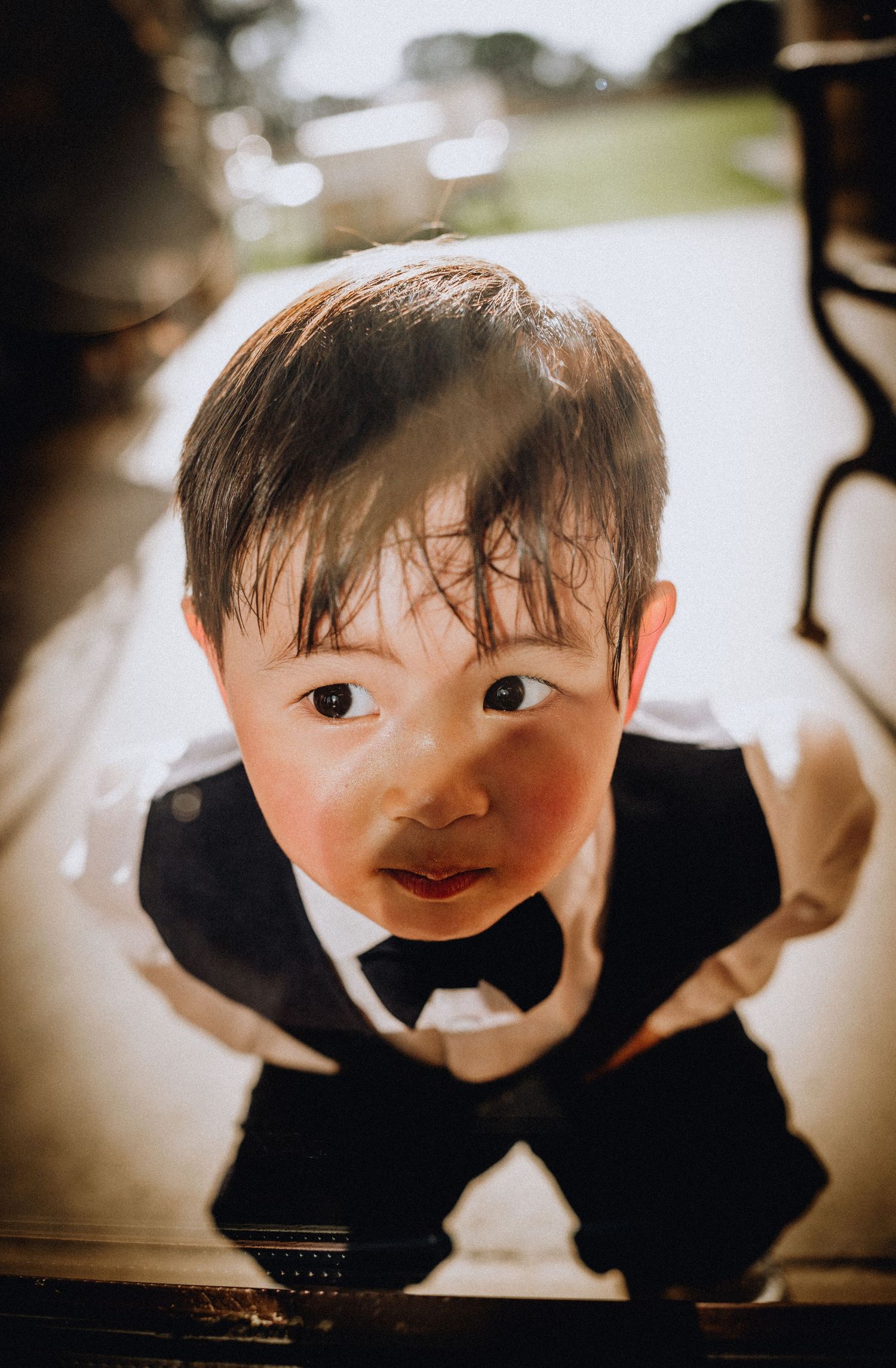 A young Asian toddler boy in a black tuxedo with bow tie looks up at the camera with curious eyes.
