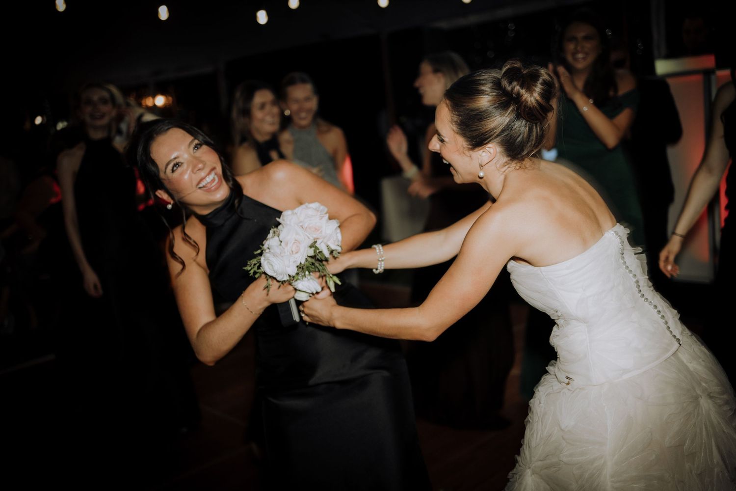 Bride in white gown handing bouquet to bridesmaid in black dress on wedding reception dance floor.