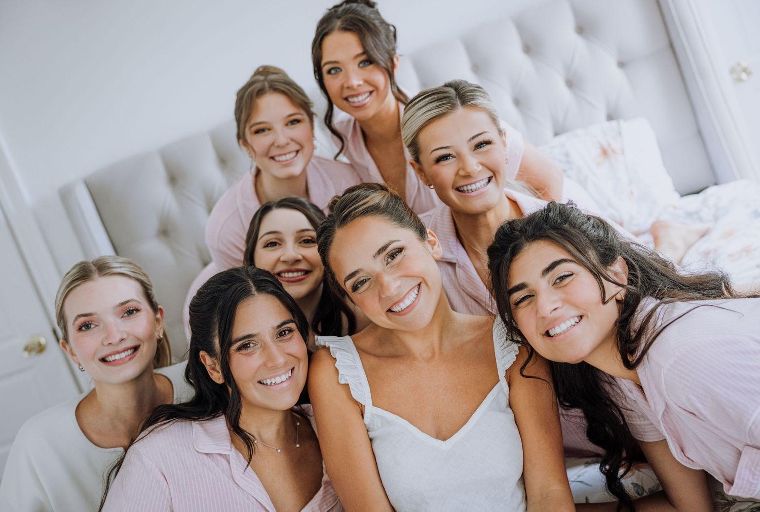 Bride in white dress smiles with bridesmaids in pink robes during wedding morning preparations.