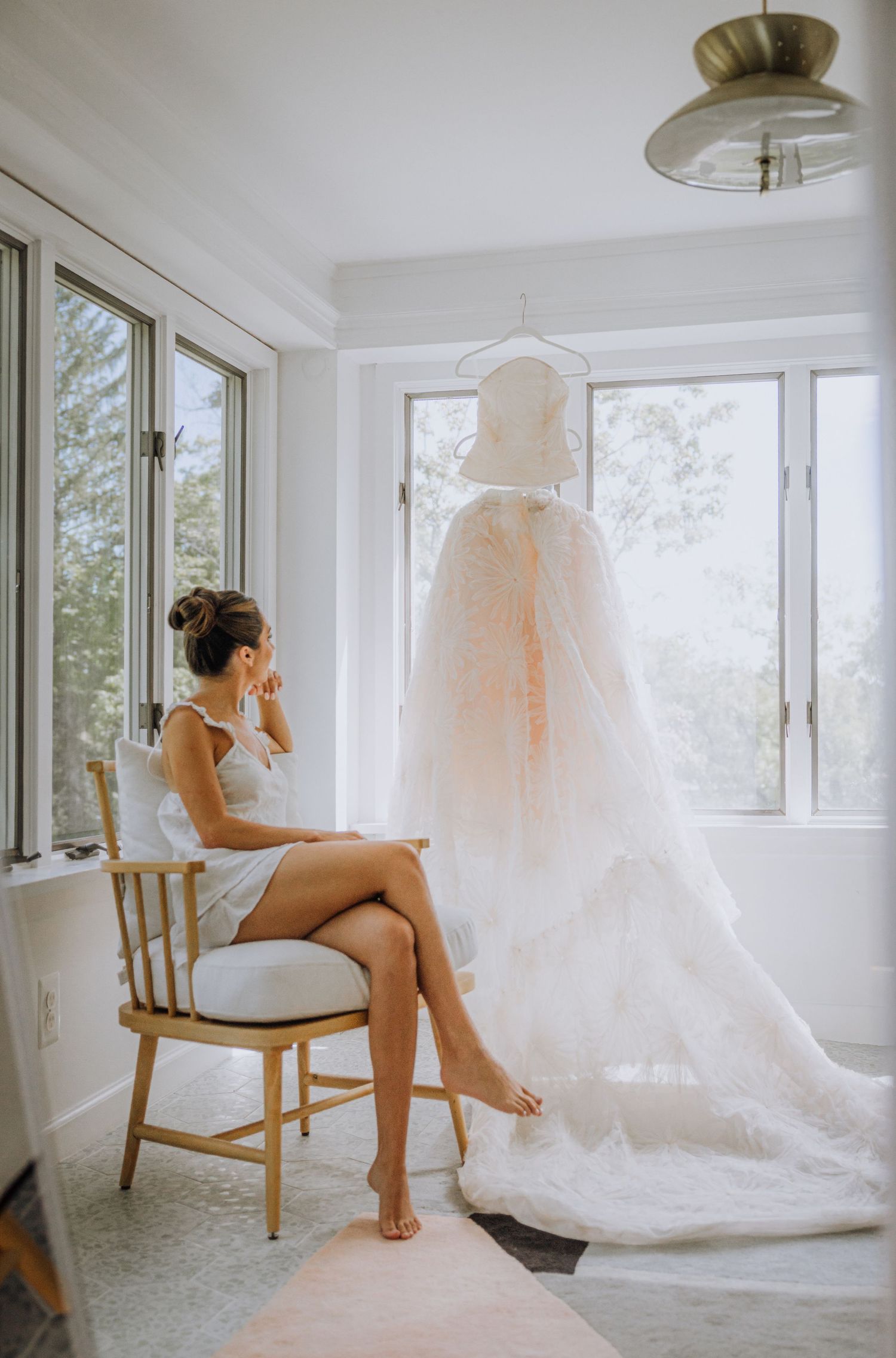 Bride in white lingerie sits on gold chair gazing at her elegant lace wedding gown hanging by sunlit windows.
