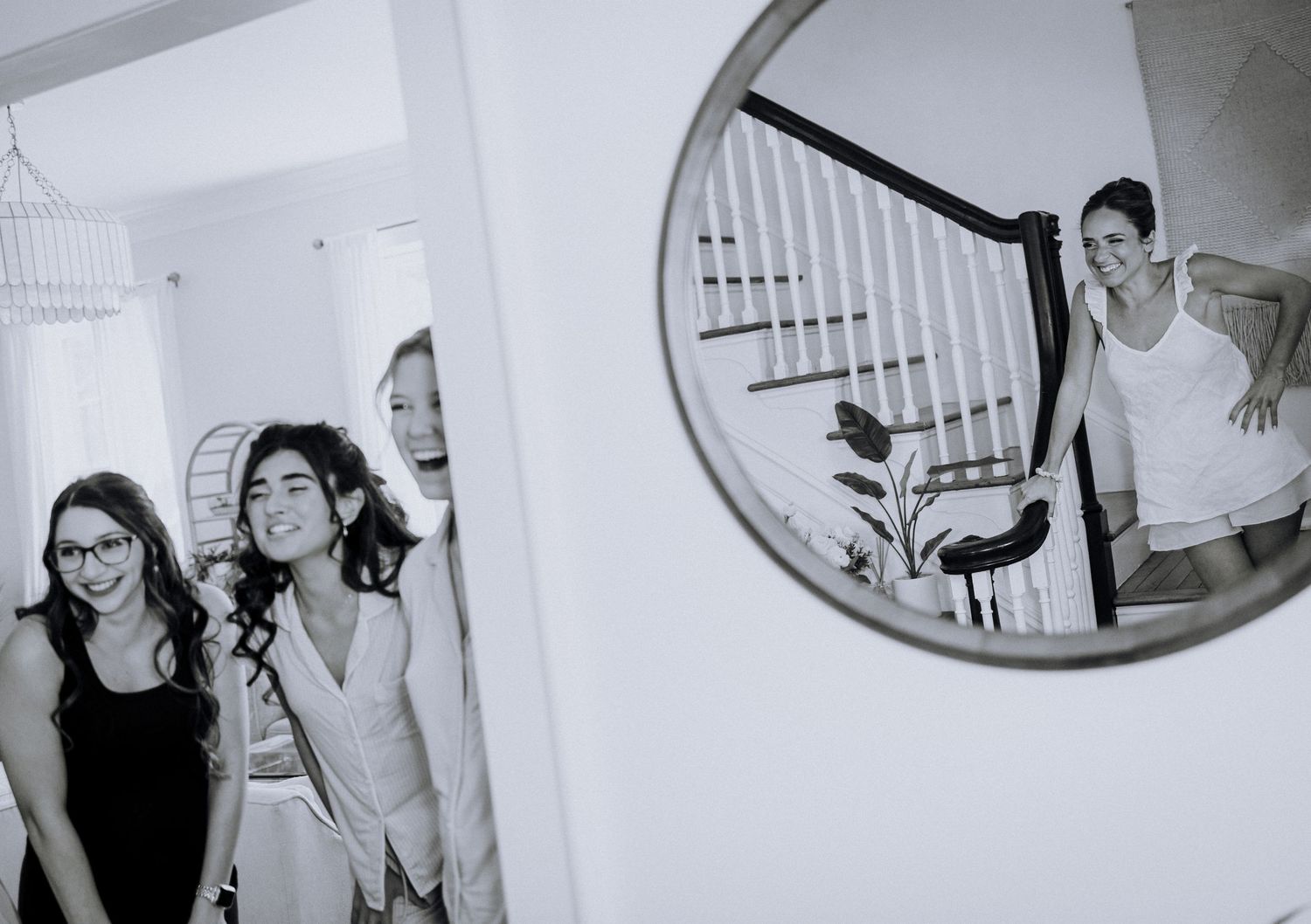 Black and white photo of three women laughing in a hallway with a man reflected in a round mirror on the wall.