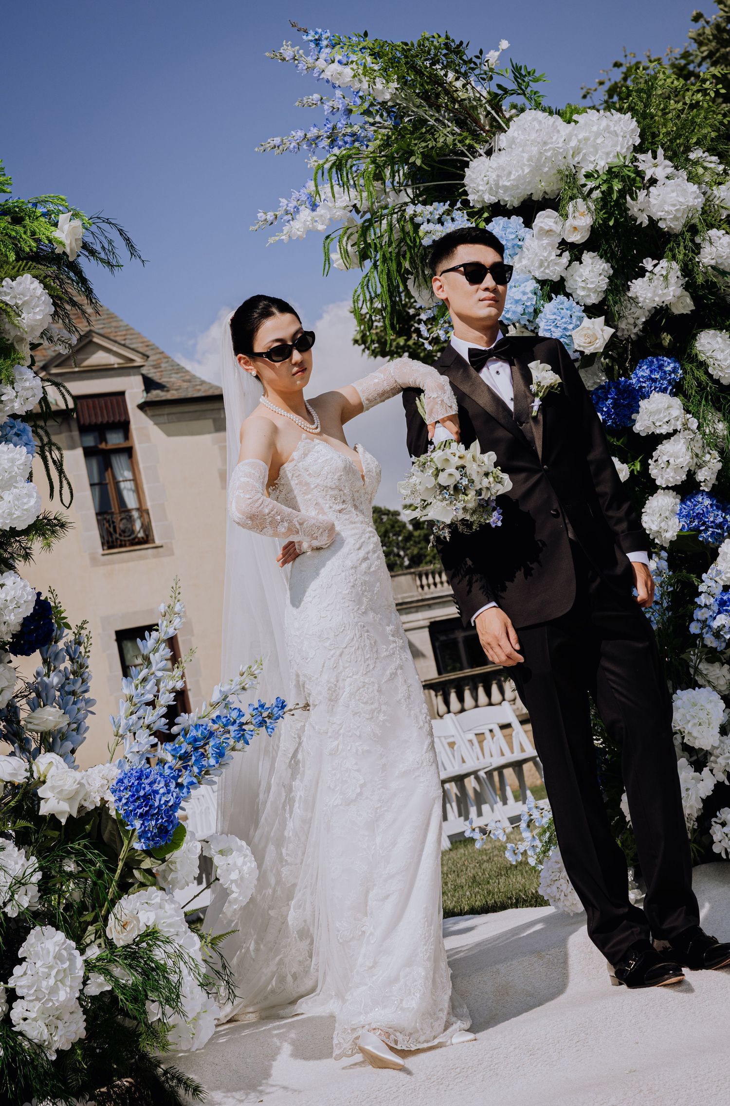 Bride in lace gown and groom in black tuxedo pose under blue and white floral arch at outdoor wedding ceremony.