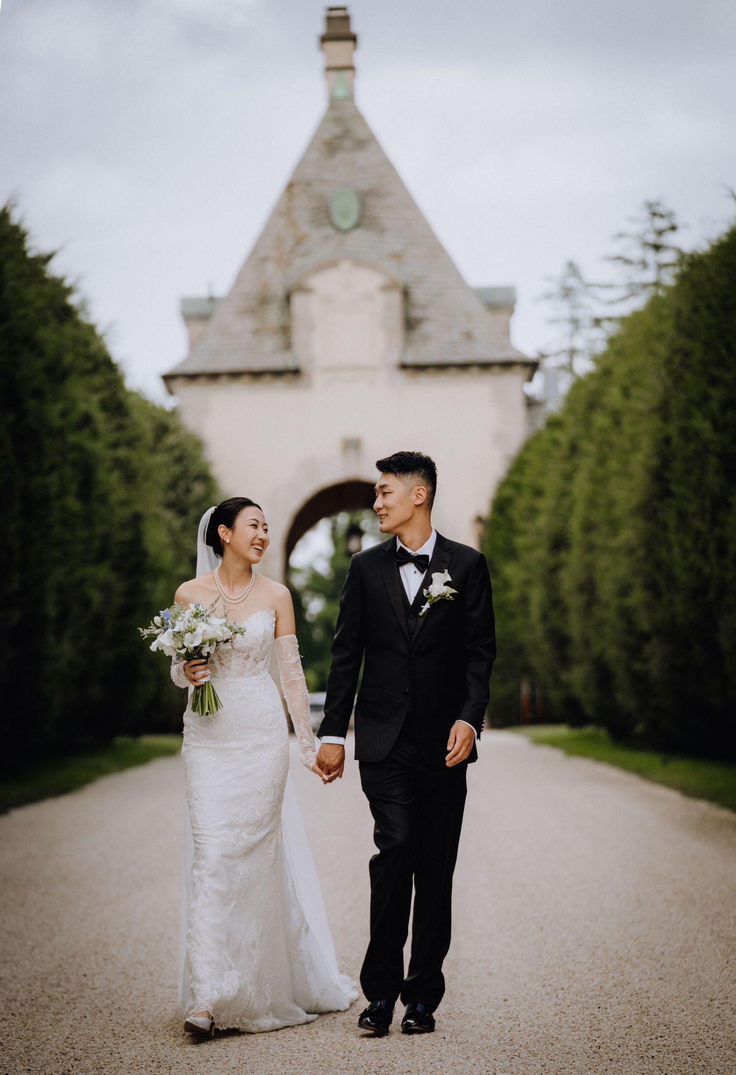 A bride and groom walk hand-in-hand along a tree-lined path before a stone archway on their wedding day.