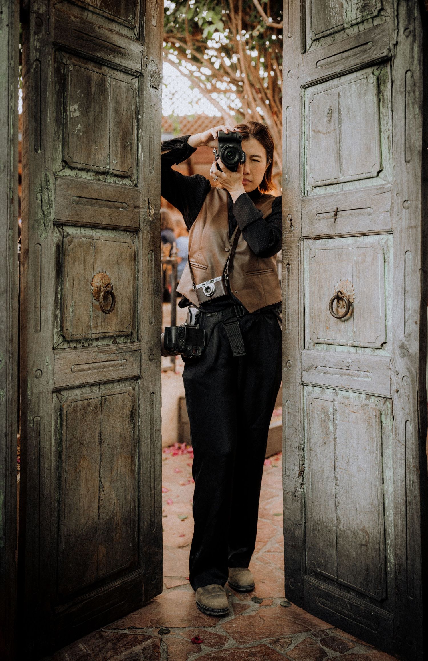 A female photographer stands in rustic wooden doorway, holding camera to face, with second camera hanging from neck.