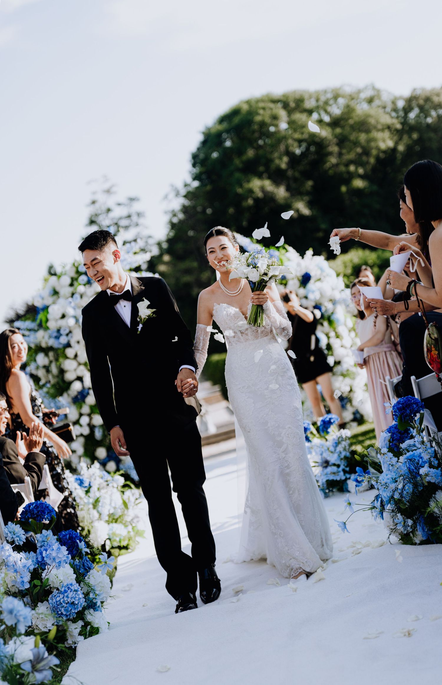 Bride and groom walk down floral aisle lined with blue hydrangeas at outdoor wedding ceremony.