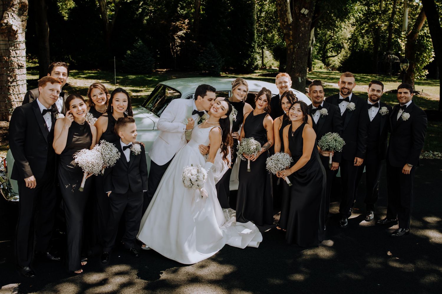 Wedding party in black attire poses with bride in white gown and groom in tuxedo outdoors near vintage car.Editorial and Cinematic Wedding p