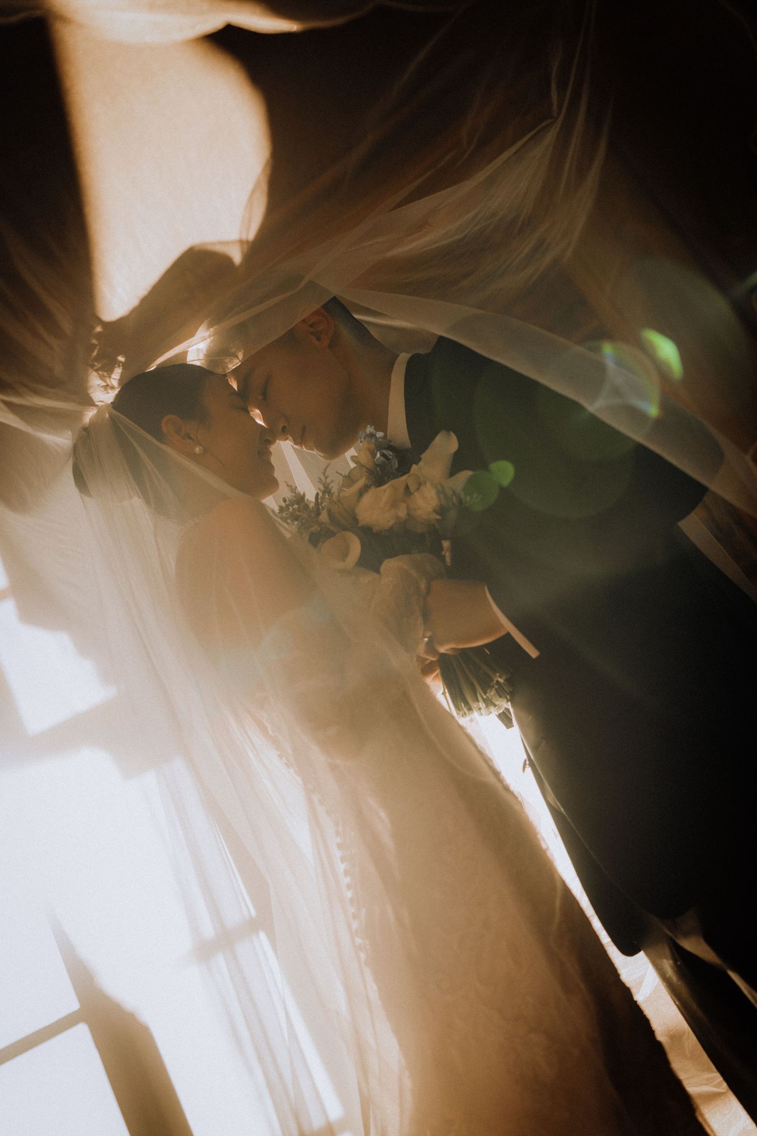 Bride and groom share a romantic kiss under a flowing veil with golden sunlight and lens flare at their wedding.