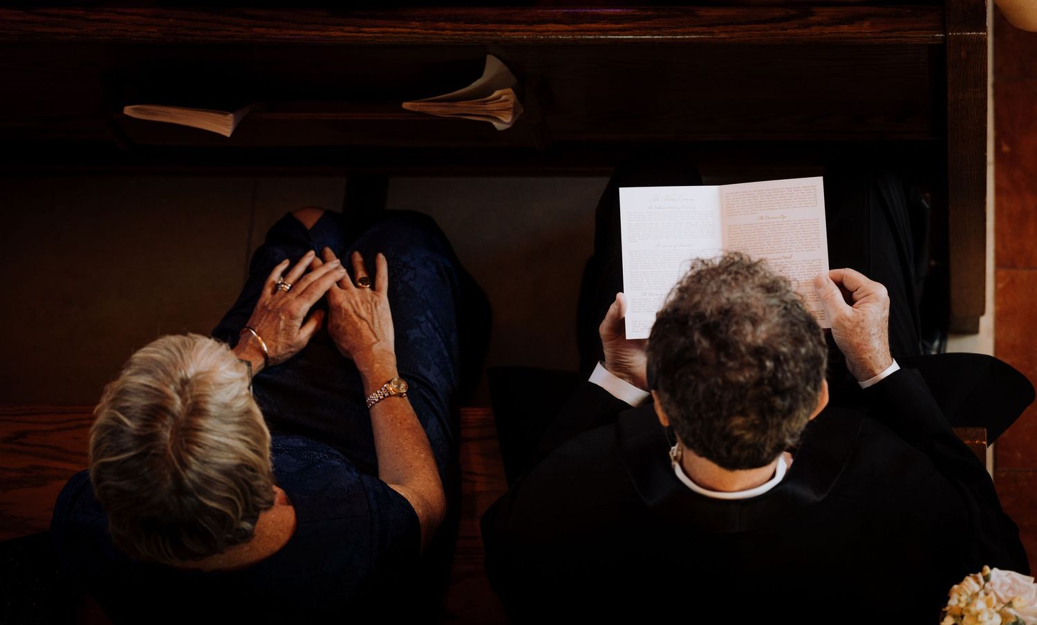 Overhead view of people seated in wooden pews, one reading a program and another with hands raised to face.