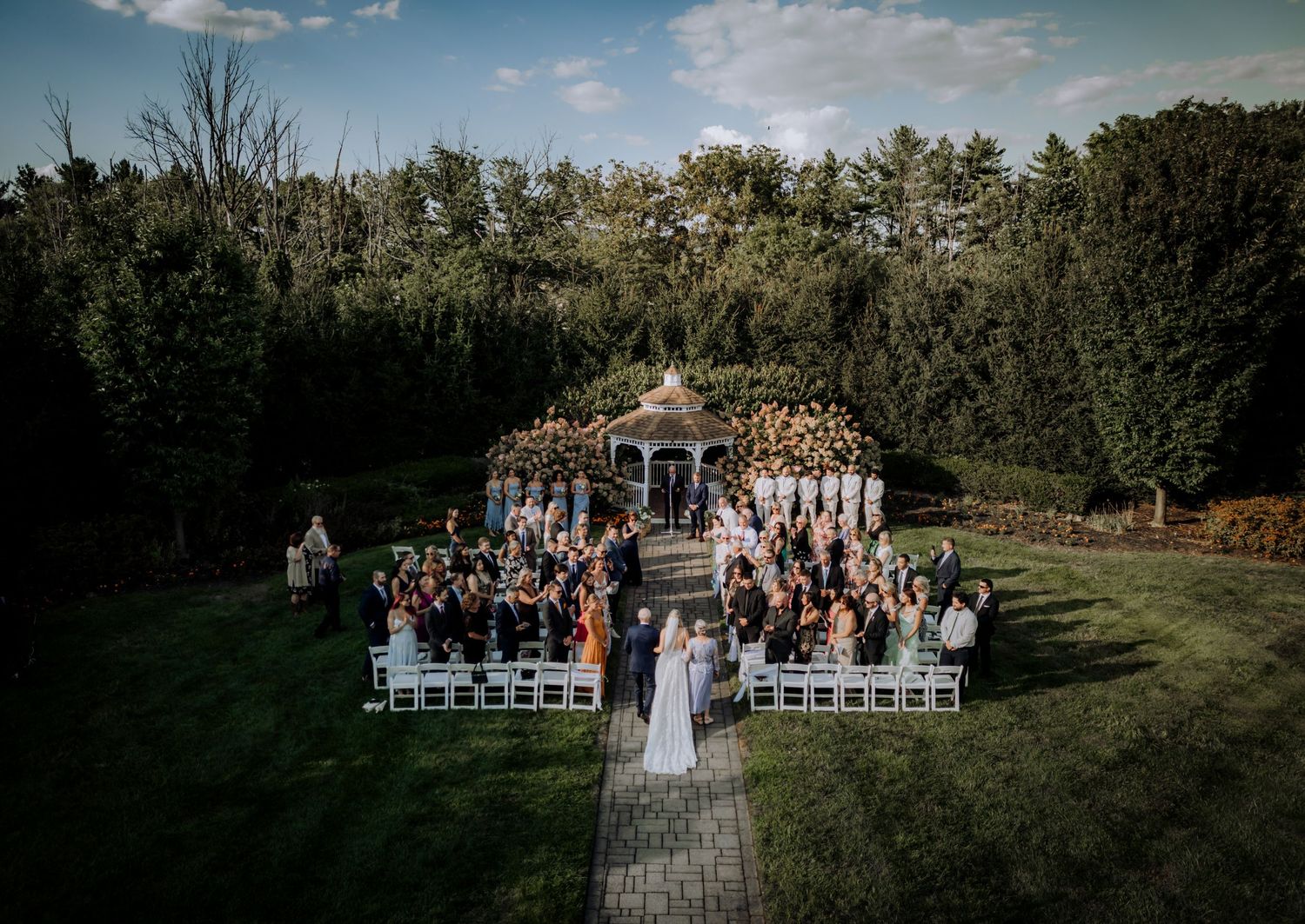 Outdoor wedding ceremony setup with white chairs and gazebo against a blue sky landscape.
