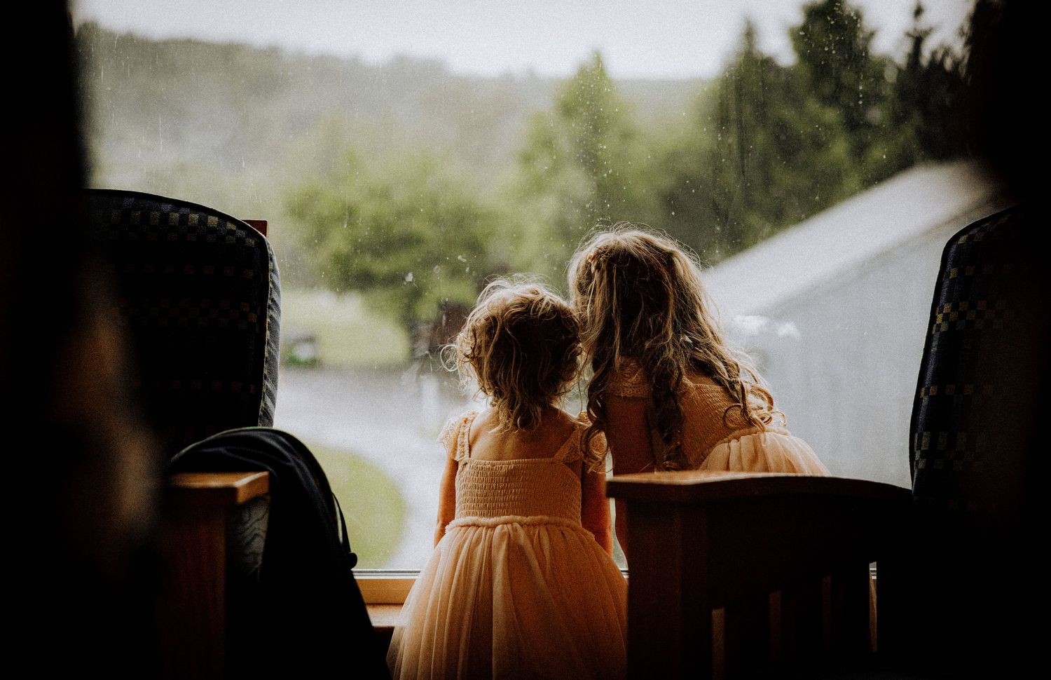 Two young girls in elegant dresses watching rain fall outside a window at a wedding venue.
