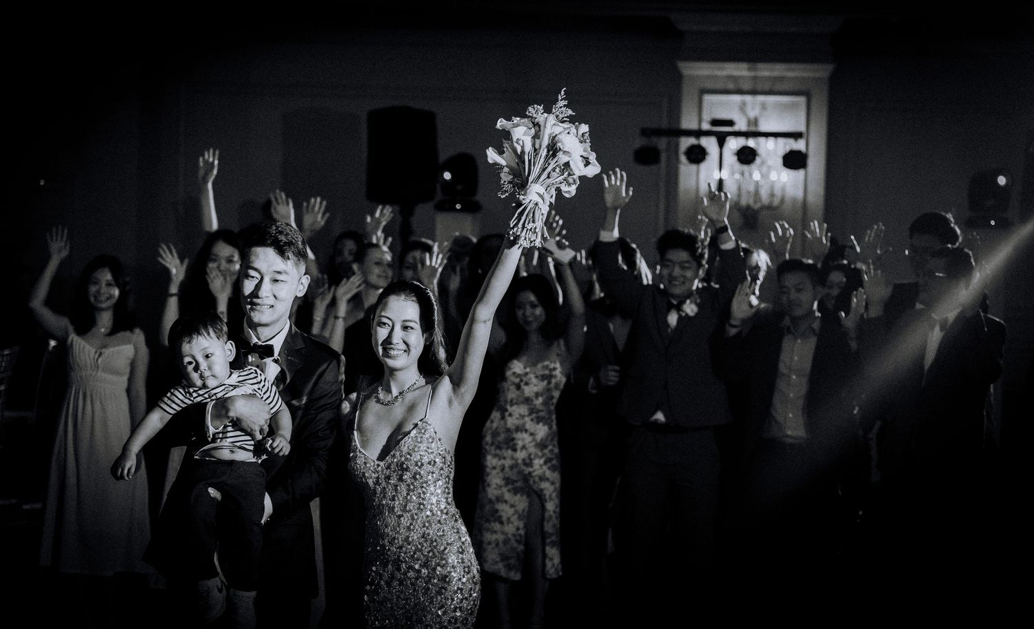 Black and white wedding reception photo of couple raising bouquet on dance floor surrounded by celebrating guests.