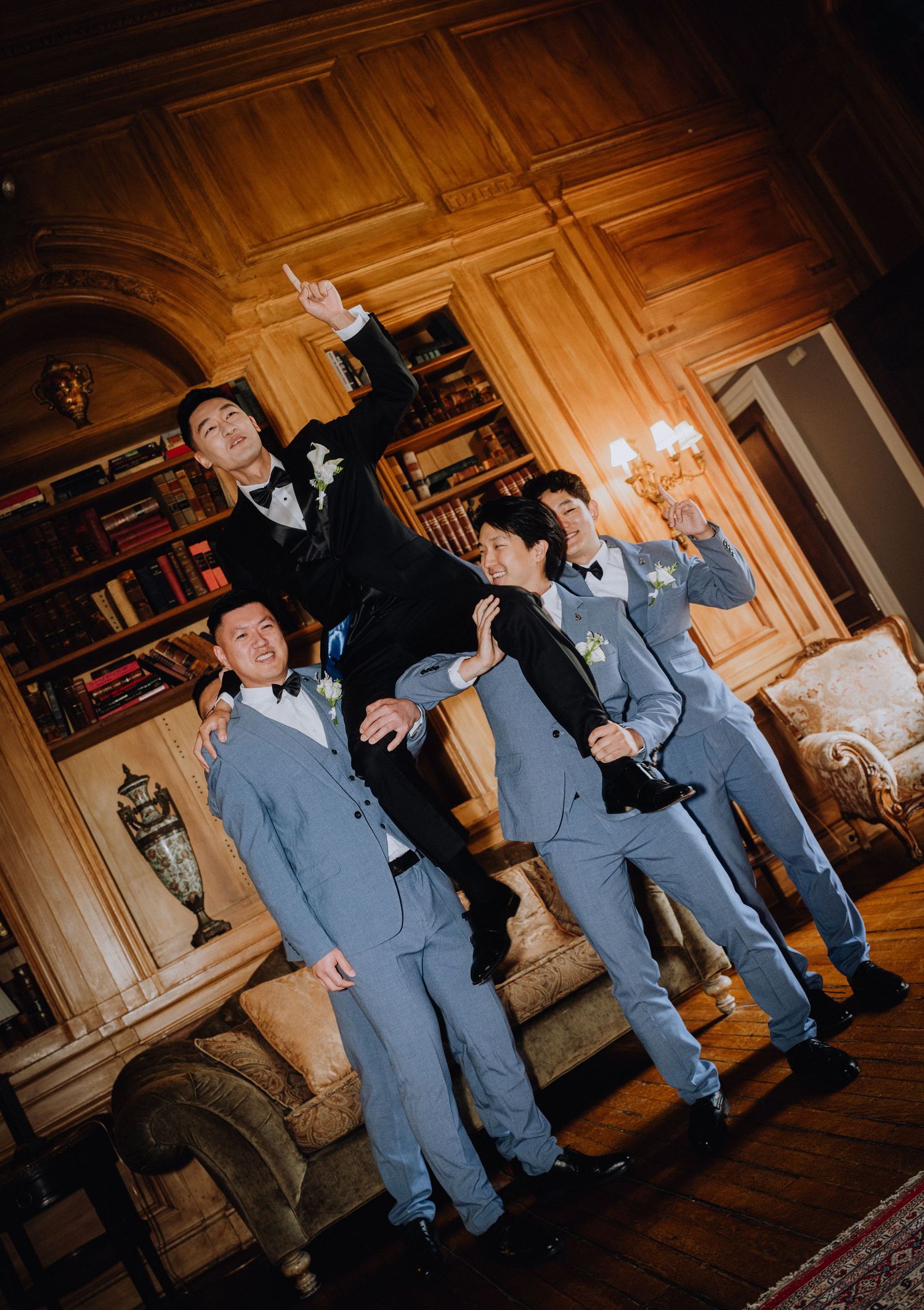 Groom in black tuxedo posing with three groomsmen in gray suits inside elegant wood-paneled room.