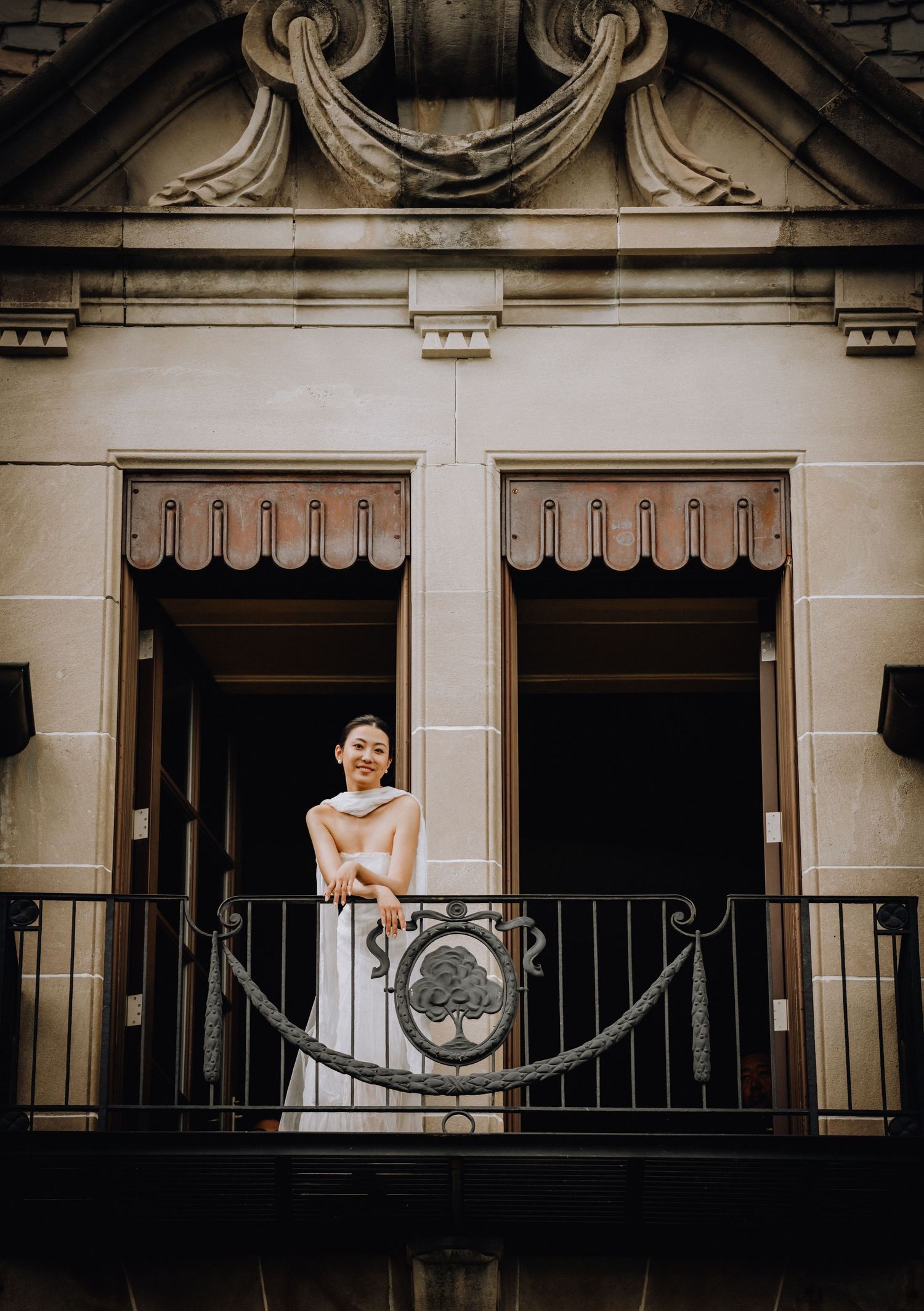 Bride in white gown smiling on ornate iron balcony of elegant stone building with decorative architecture.