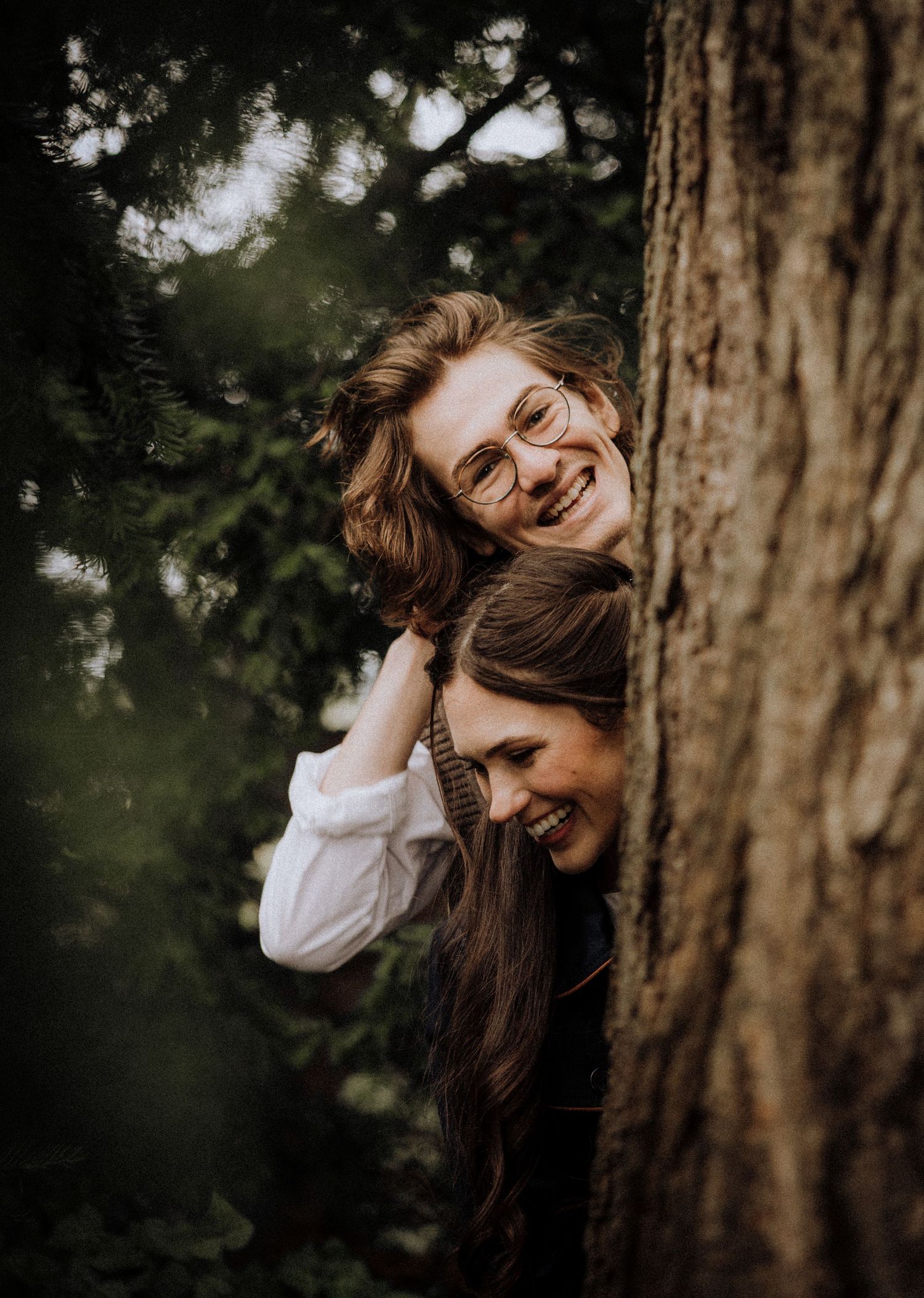 Playful moment captured between couple peeking around large tree trunk in forest setting.