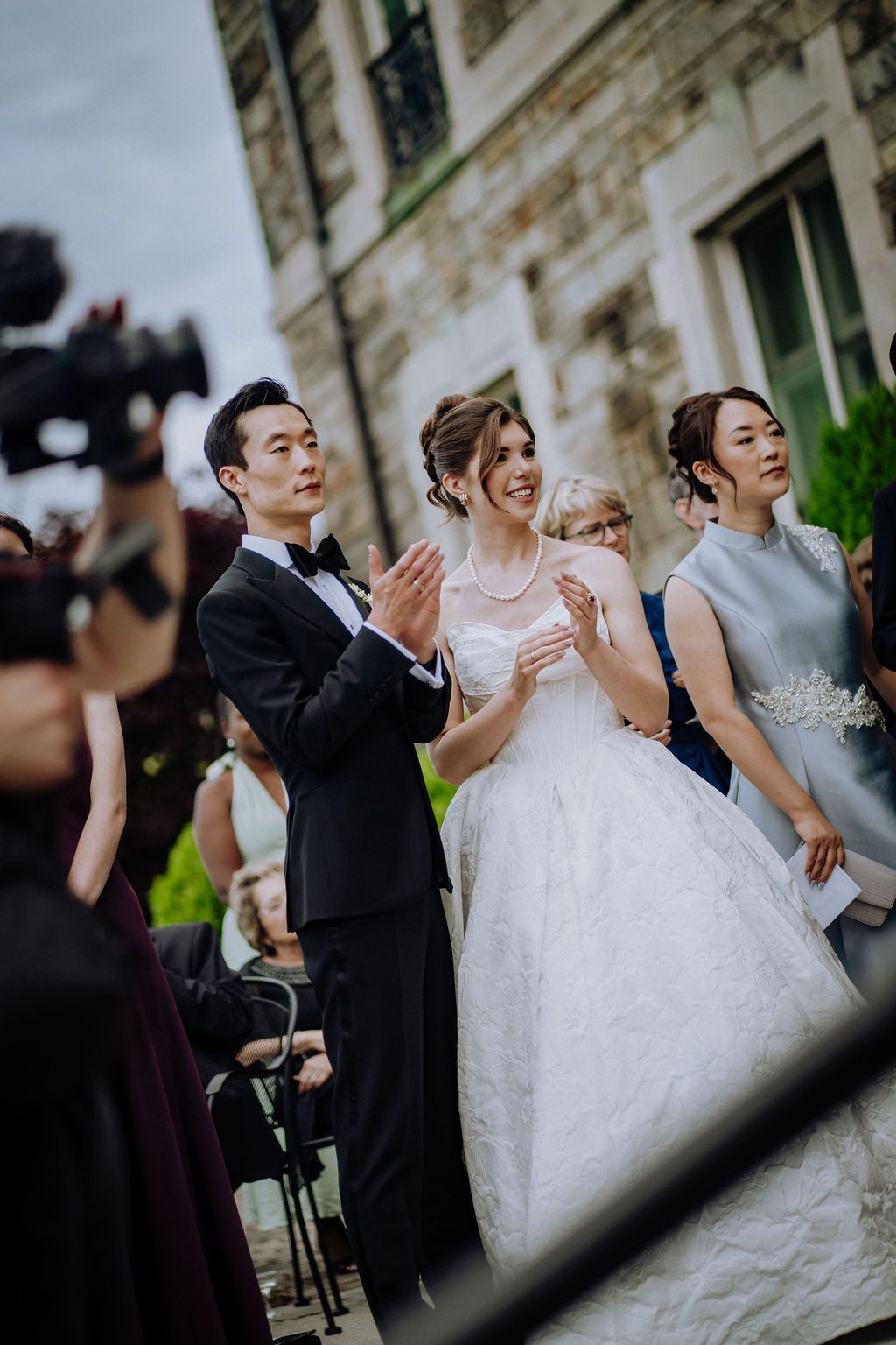 Bride and groom clapping outdoors at wedding ceremony with guests and videographer nearby.