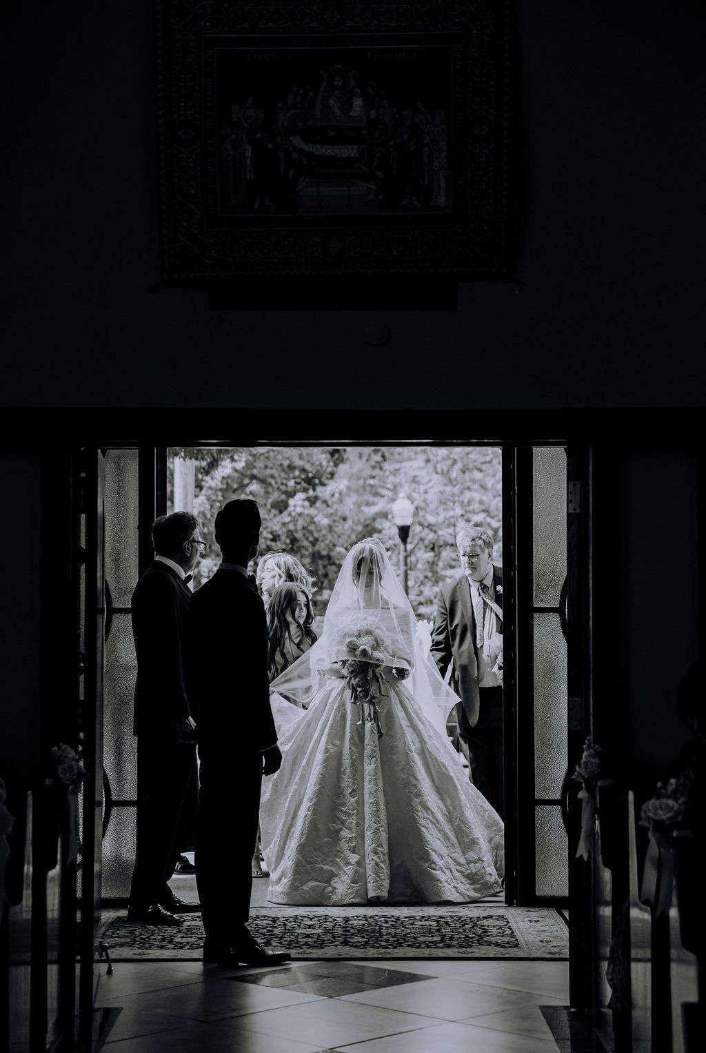 Silhouette of bride in elegant gown standing at church doors with wedding party in sunlight.
