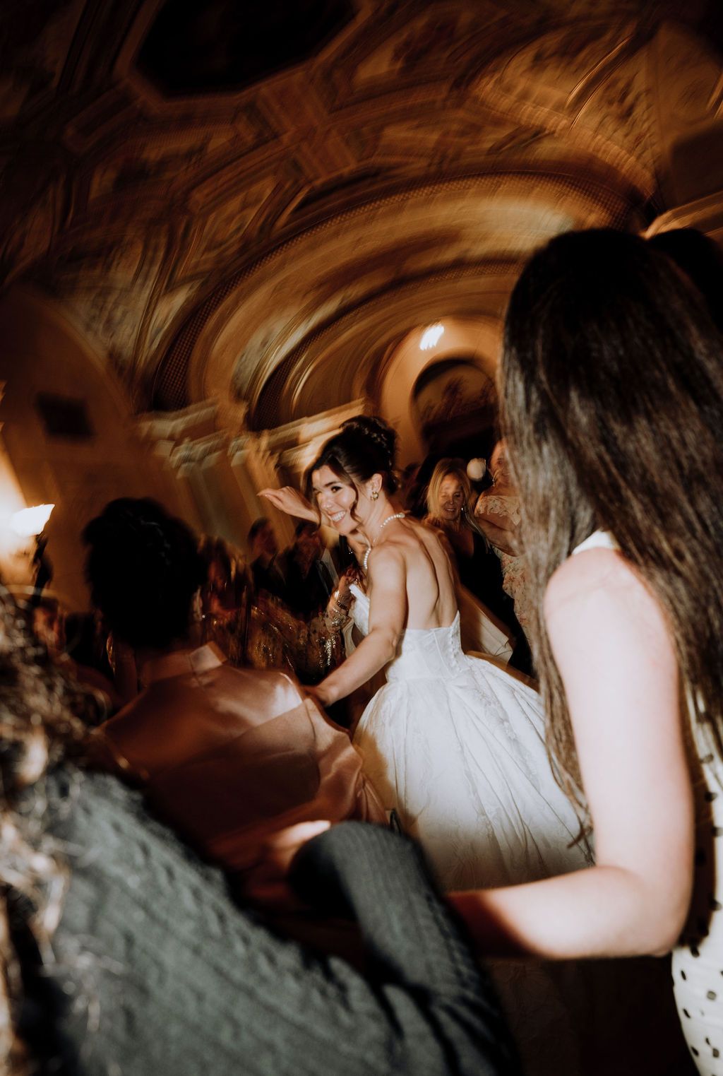 Bride in white tulle gown dancing joyfully with guests in an ornate vaulted ceiling venue.