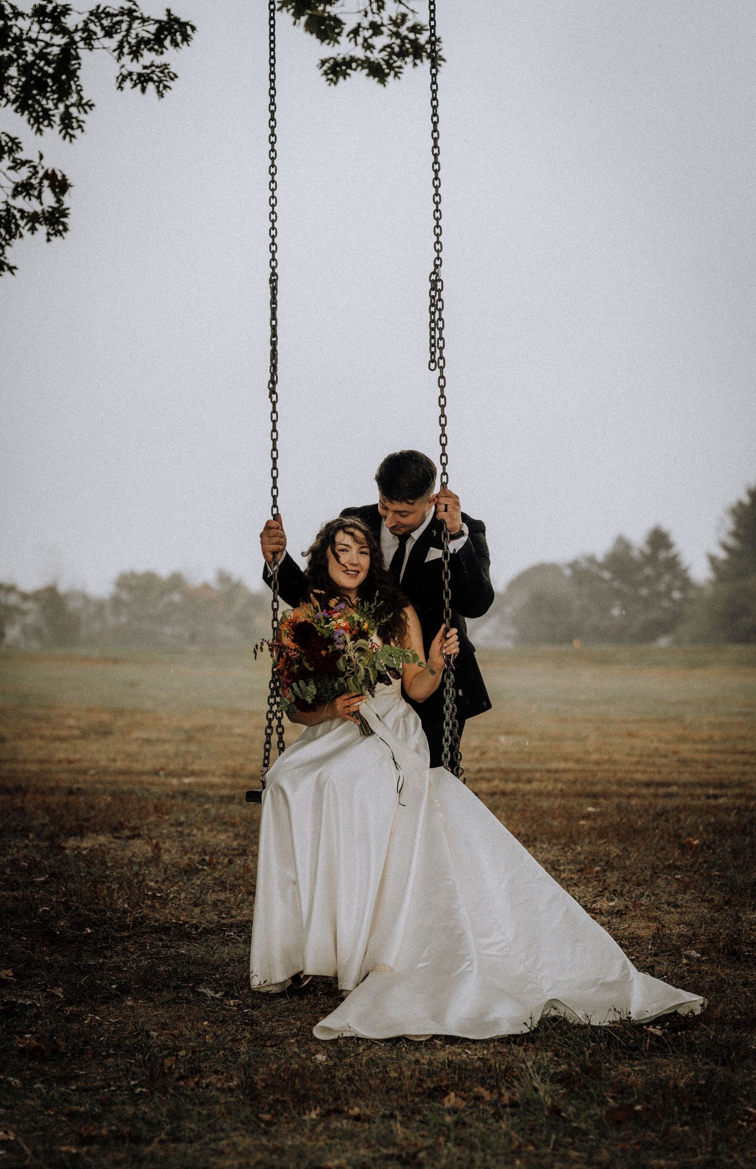 Newlyweds share an intimate moment on a swing in a misty field during their wedding photoshoot.